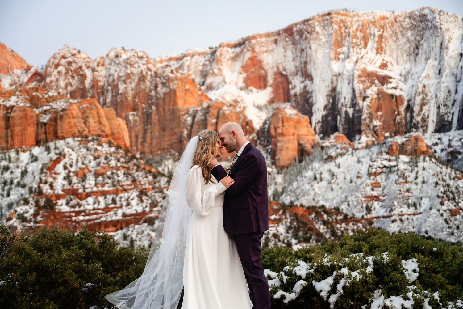 Winter wedding portraits against dramatic snow-dusted red rock cliffs at sunset.
