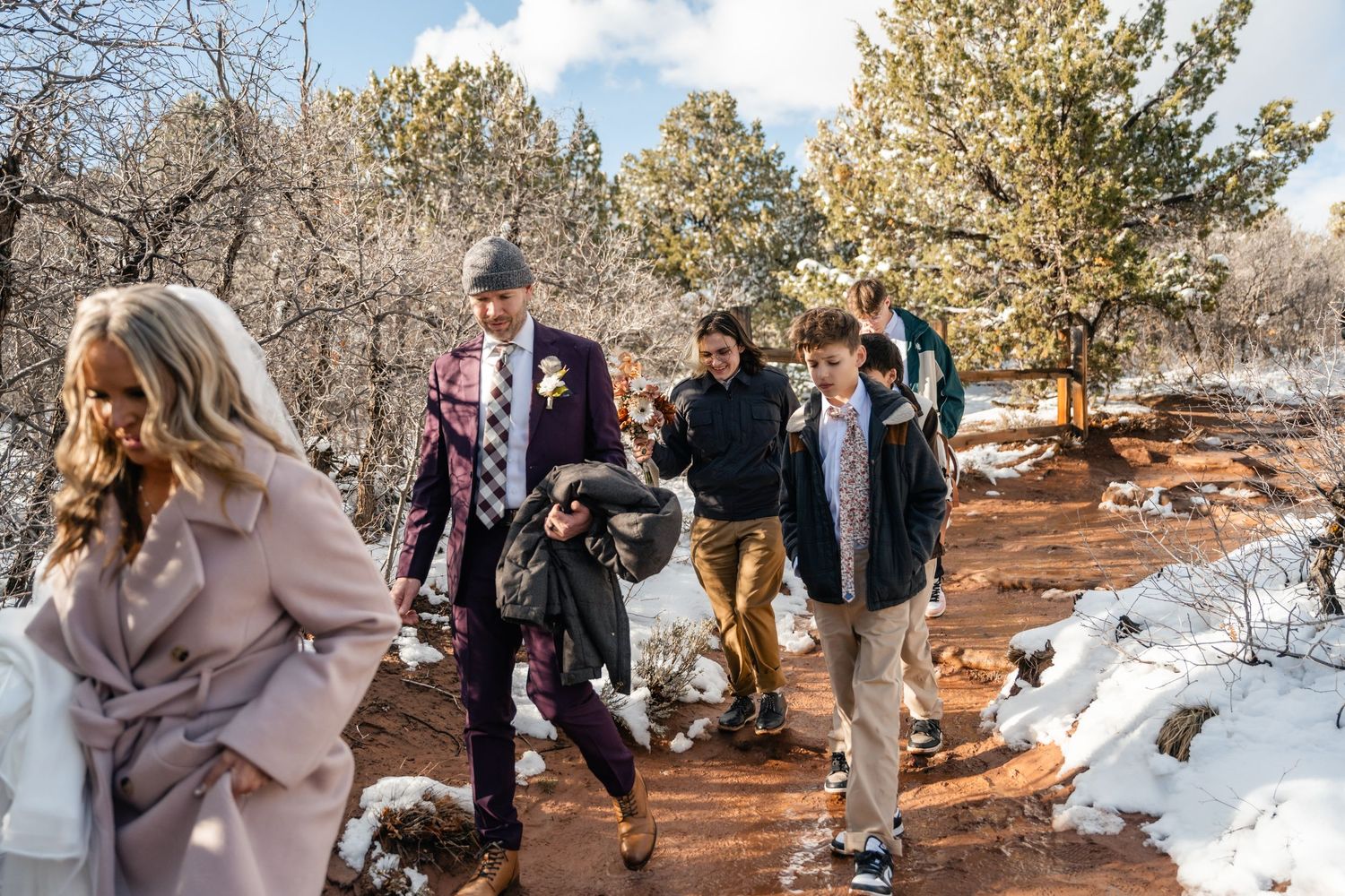 Wedding party walking along a snowy trail with red rock formations in background.