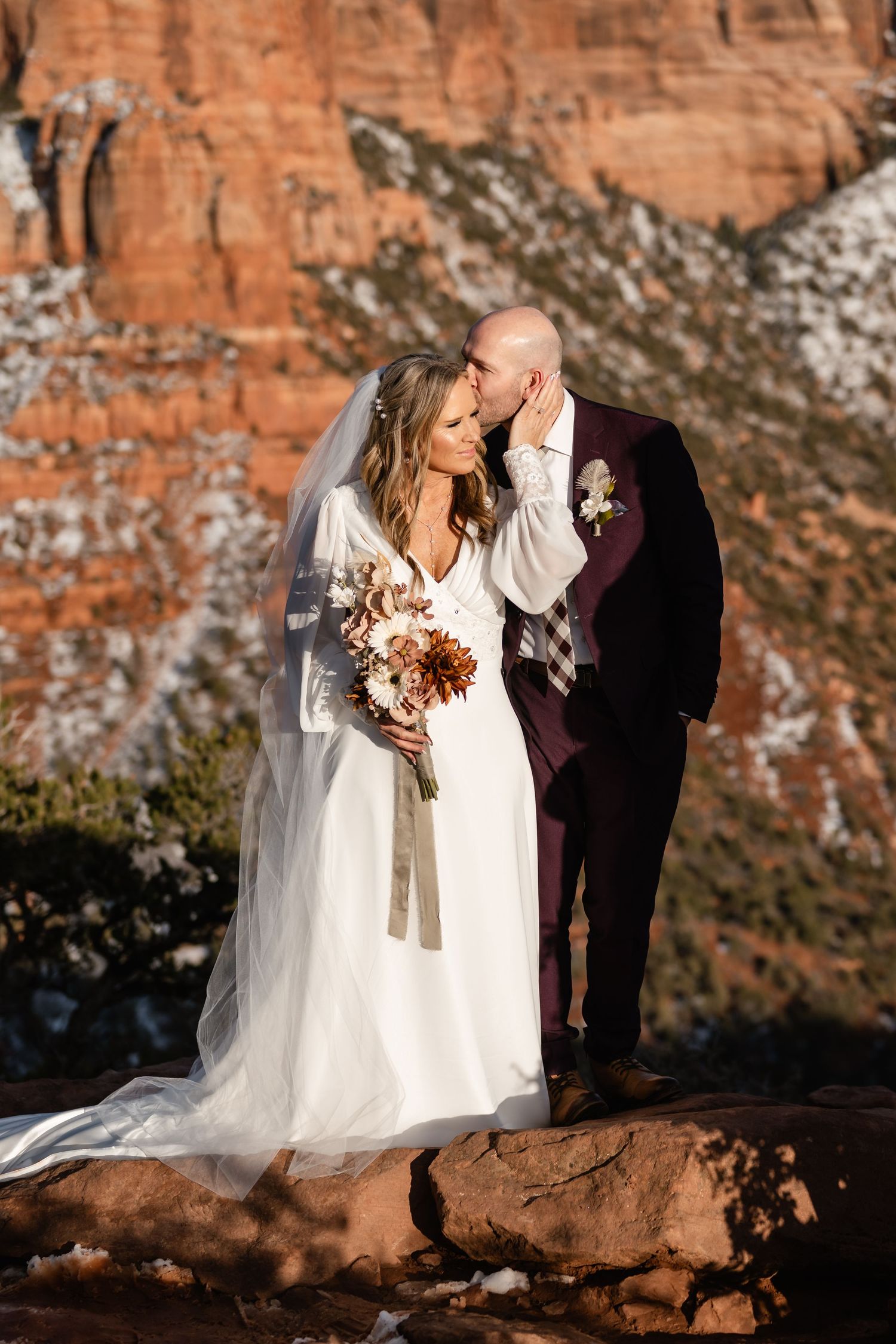 Wedding couple poses on red rocks during sunset with snow-dusted cliffs in background.