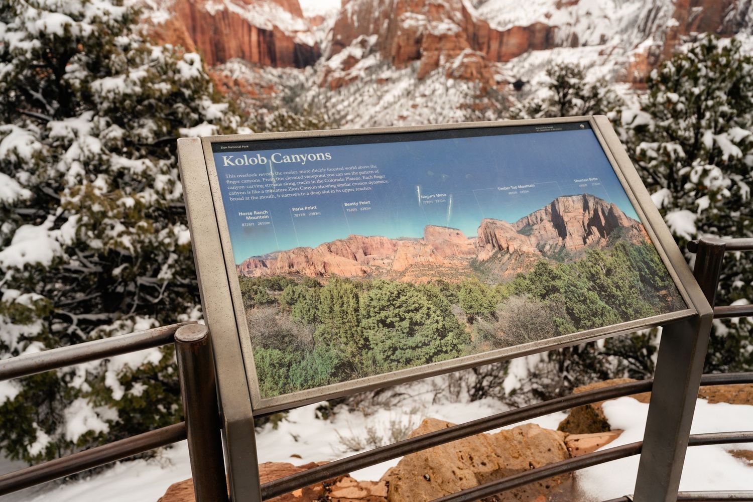 Snowy winter landscape of red rock formations and informational signs at a scenic overlook in a national park.