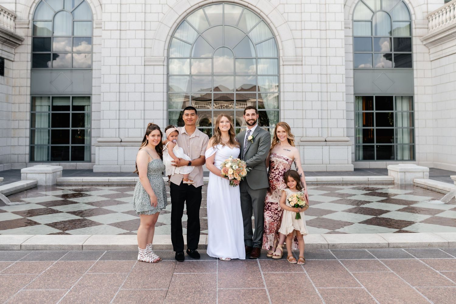 Wedding party poses together in front of elegant white stone building with arched windows and marble columns.