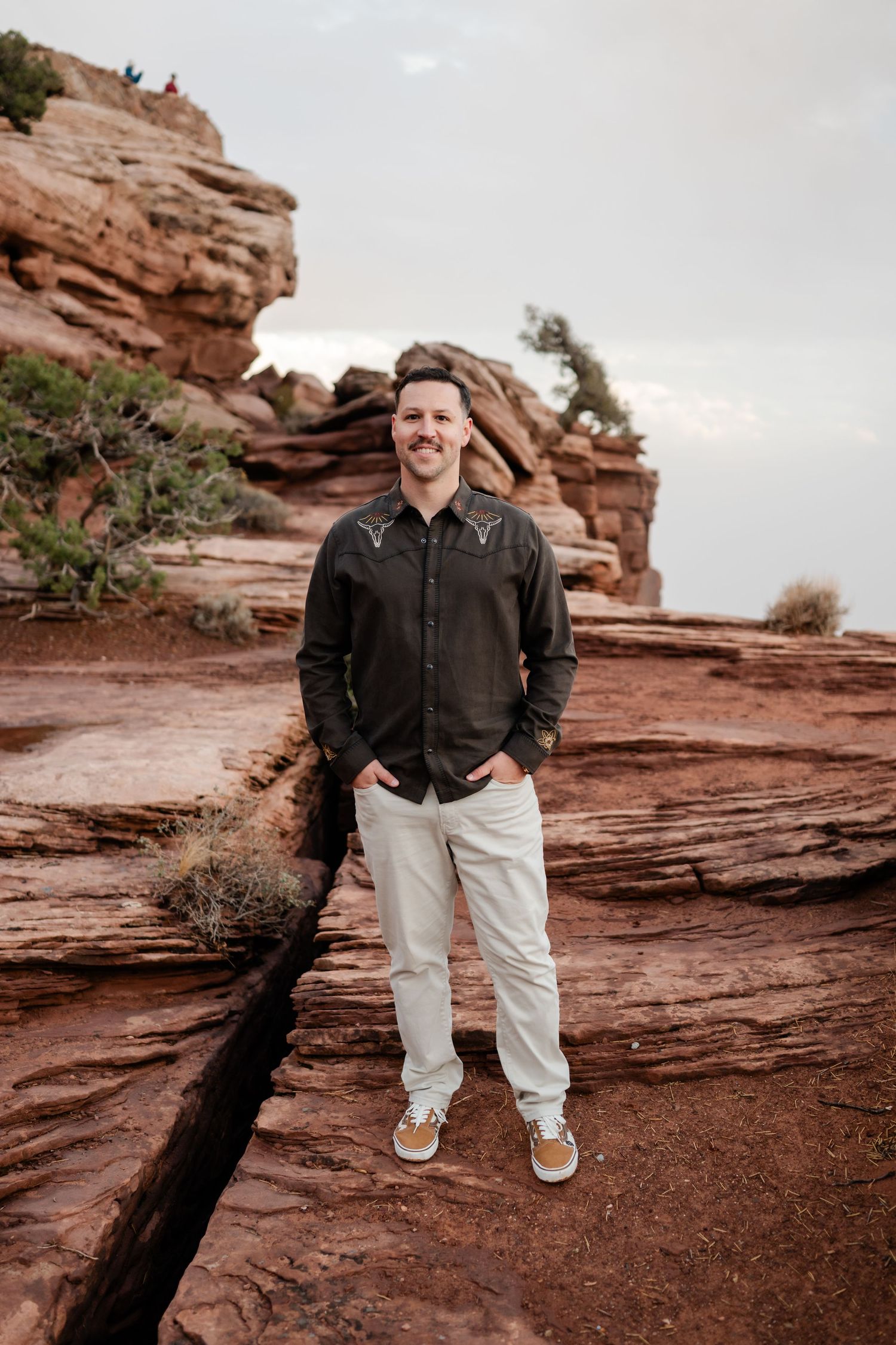 A person stands casually on red rock formations wearing a dark shirt and light pants.