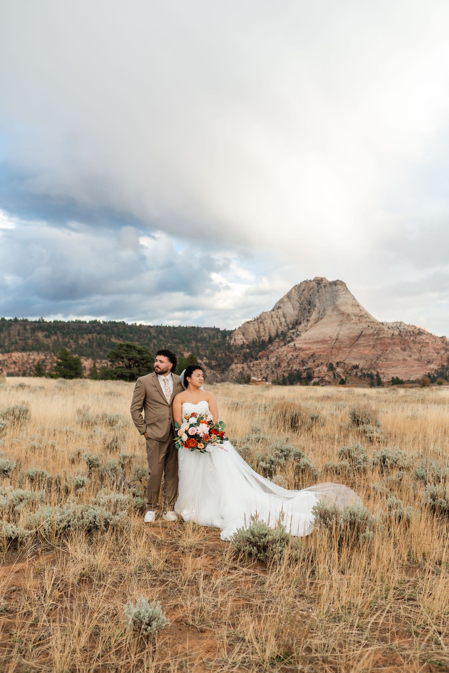 Wedding couple shares romantic moment in field with red rock formations in background.