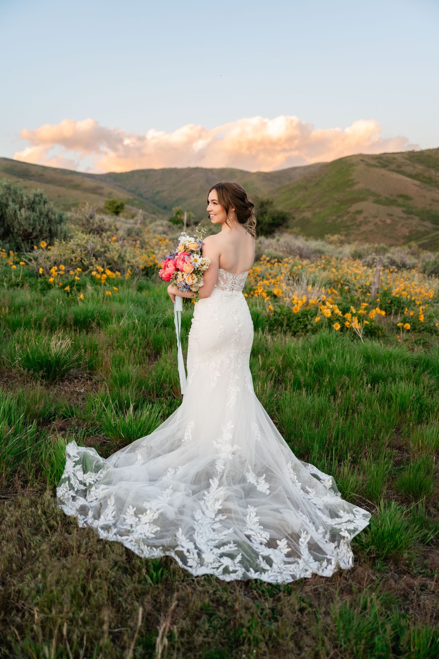 Bride with flowing lace dress stands among wildflowers during golden hour mountain photoshoot.