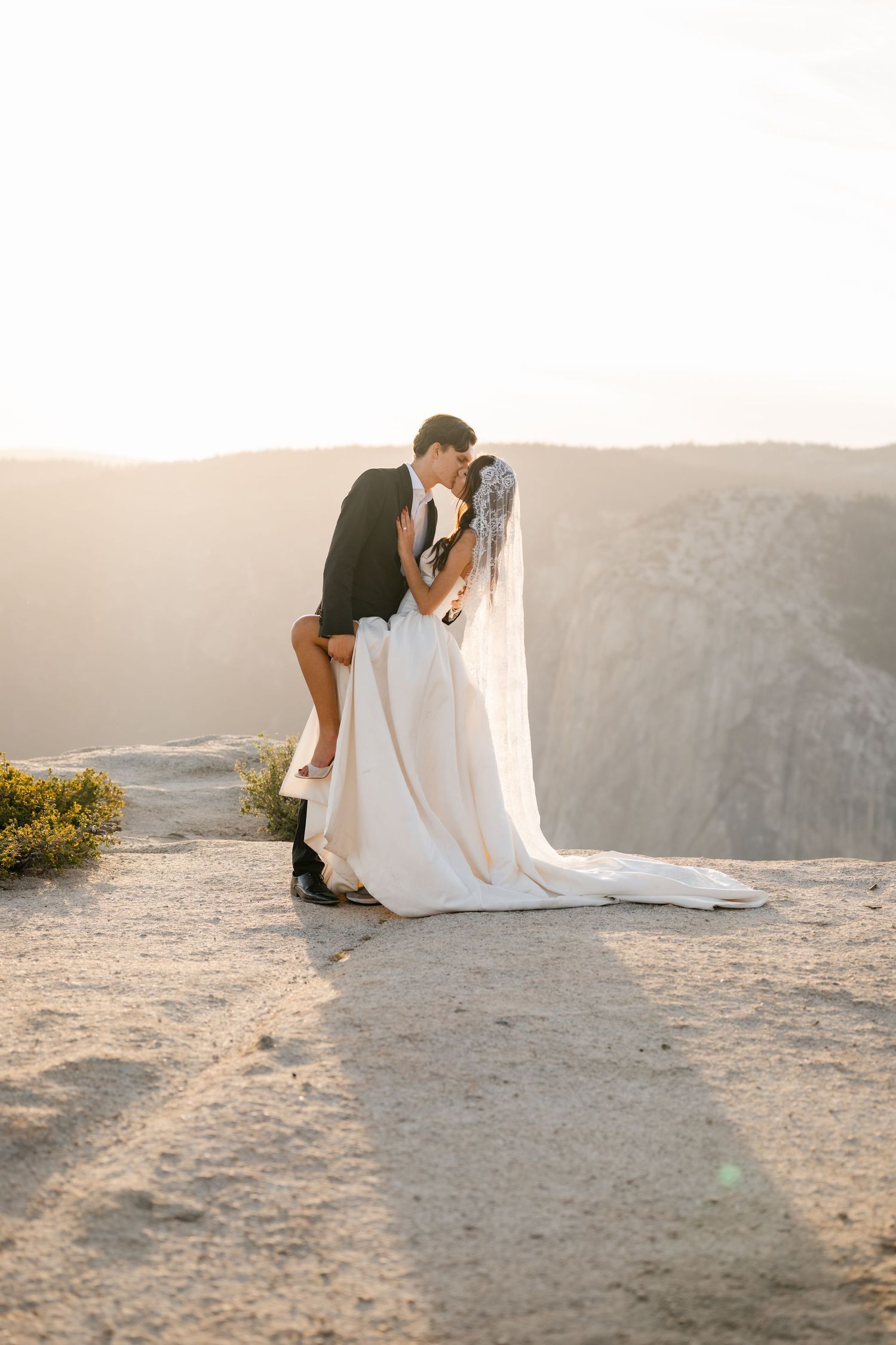 Groom dipping bride for a romantic kiss on a mountain cliff at golden hour.