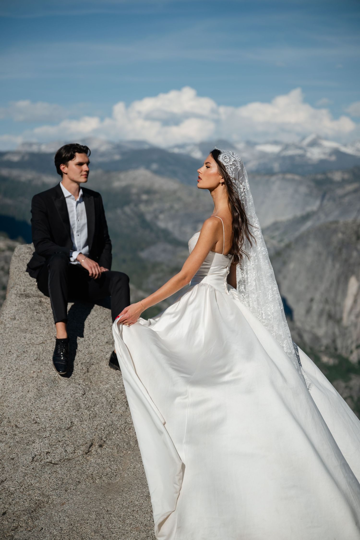 Bride in flowing white gown and veiled headpiece poses with groom on rocky Glacier Point overlook at Yosemite.