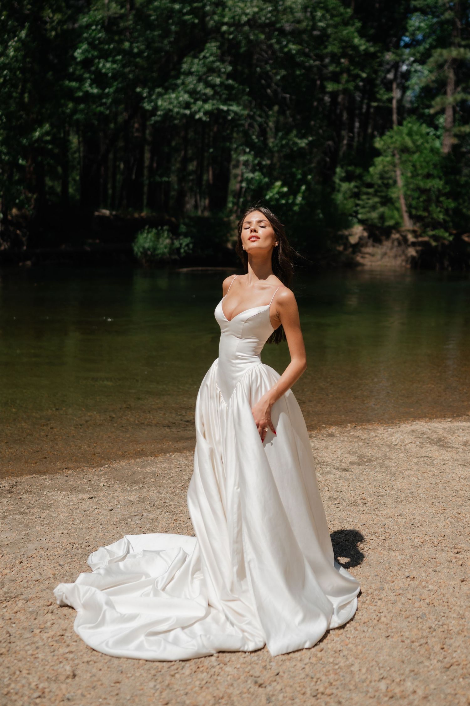 Bride in strapless white satin ballgown with cathedral train poses by a serene forest river on sandy shore of Cathedral Beach.