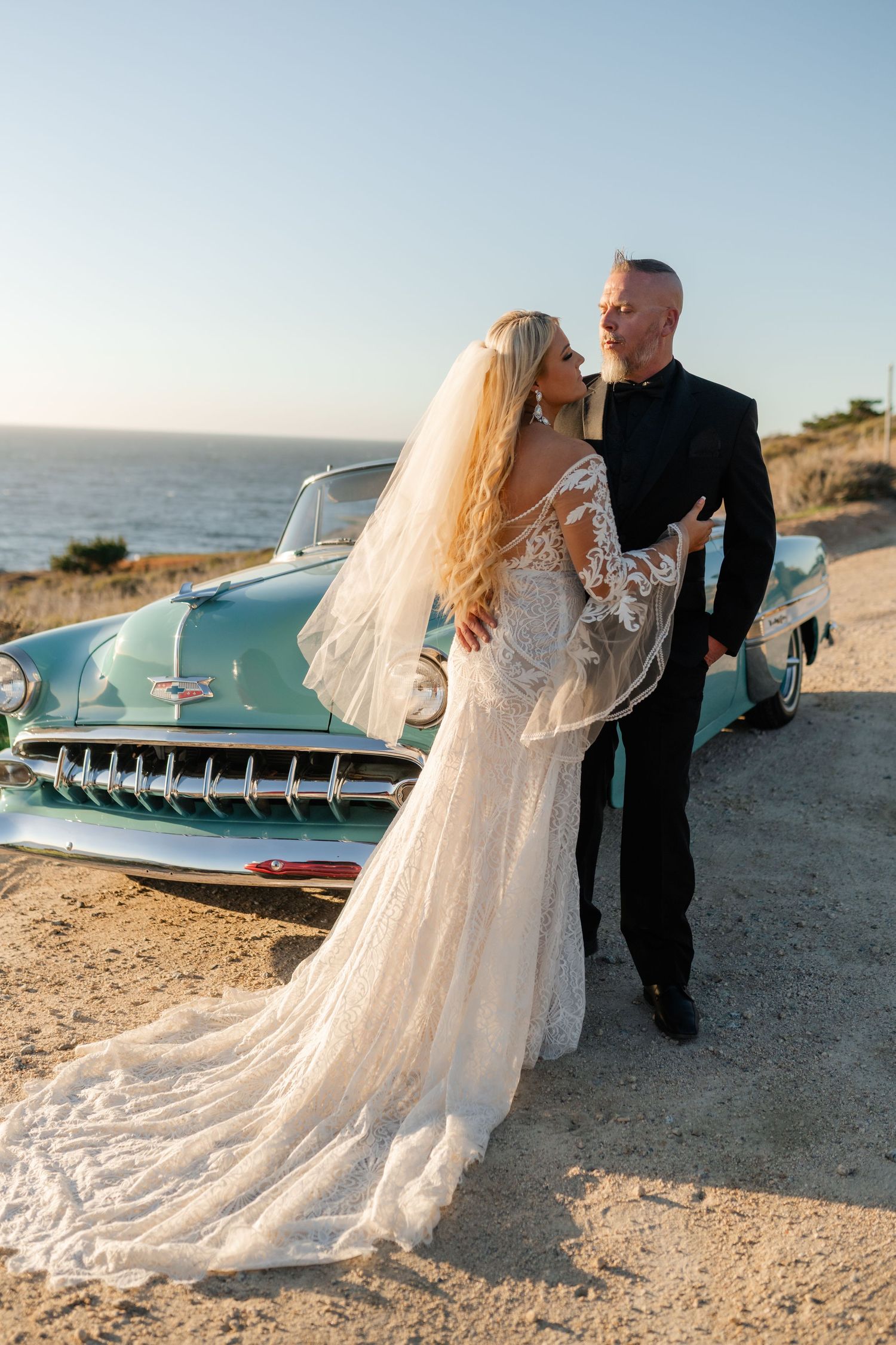 Wedding couple poses next to a vintage turquoise car on a coastal beach setting.
