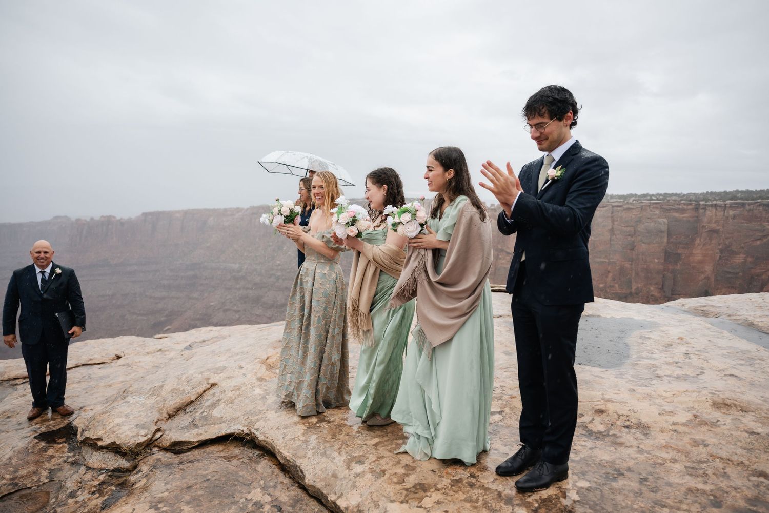 Wedding party members clap during ceremony on rocky cliff overlooking canyon.