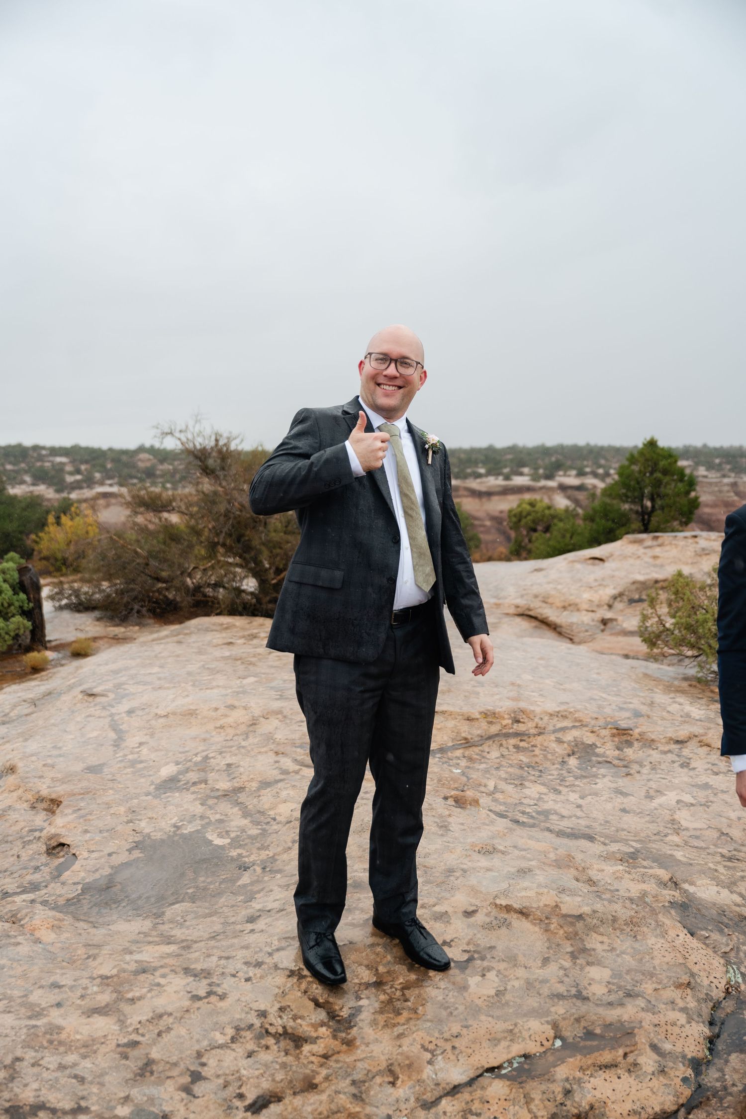 Person in dark suit giving thumbs up while standing on rocky terrain.