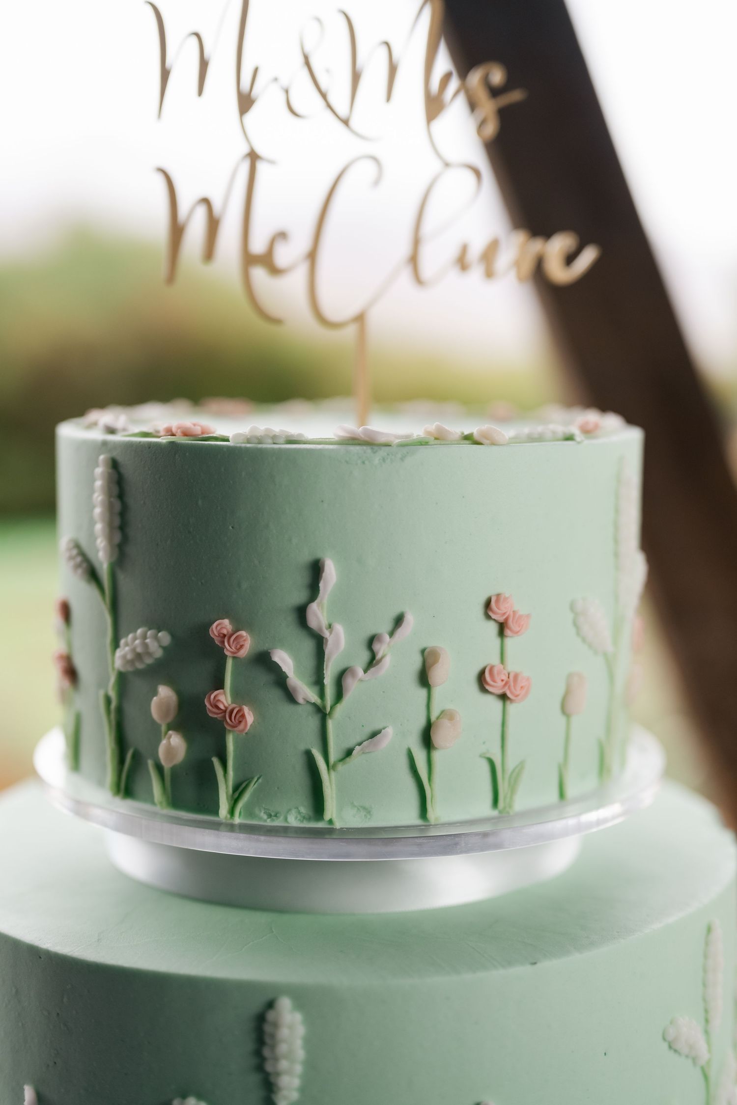 Close-up of mint green wedding cake with hand-painted floral details and gold calligraphy topper.