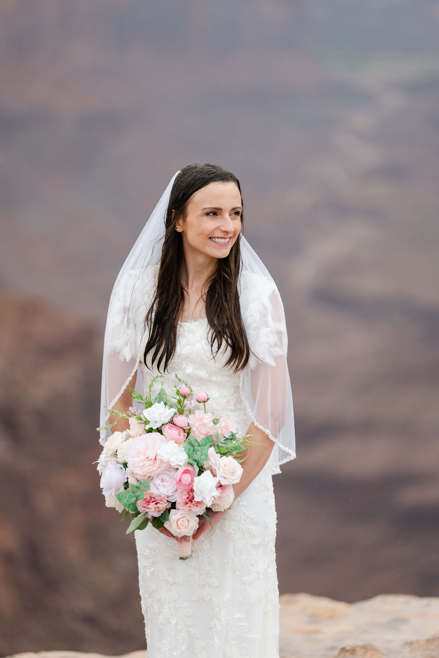 A bride in a white lace dress holds a pink and white bouquet against a desert canyon backdrop.