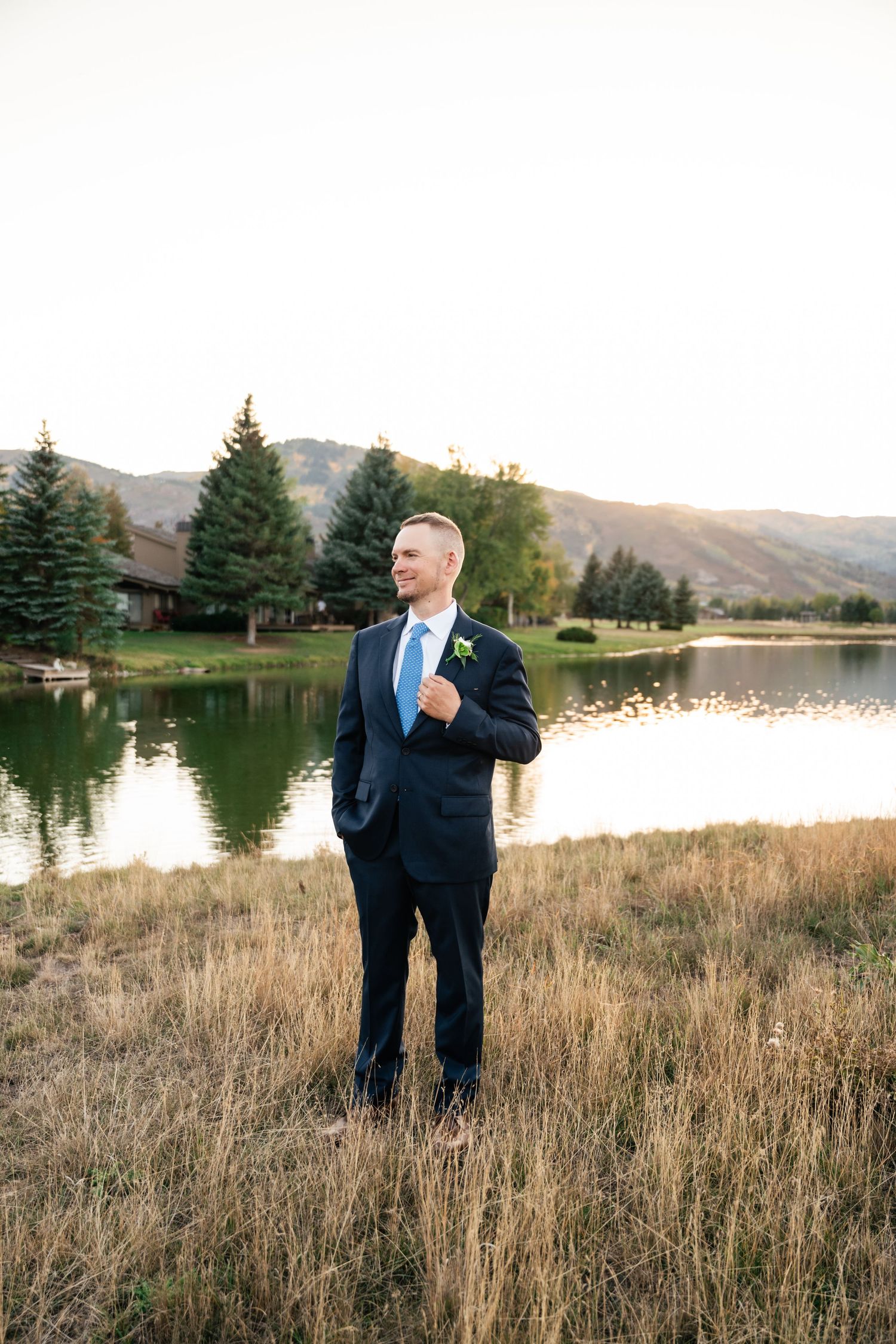 Groom in navy suit stands in field with mountain lake backdrop.
