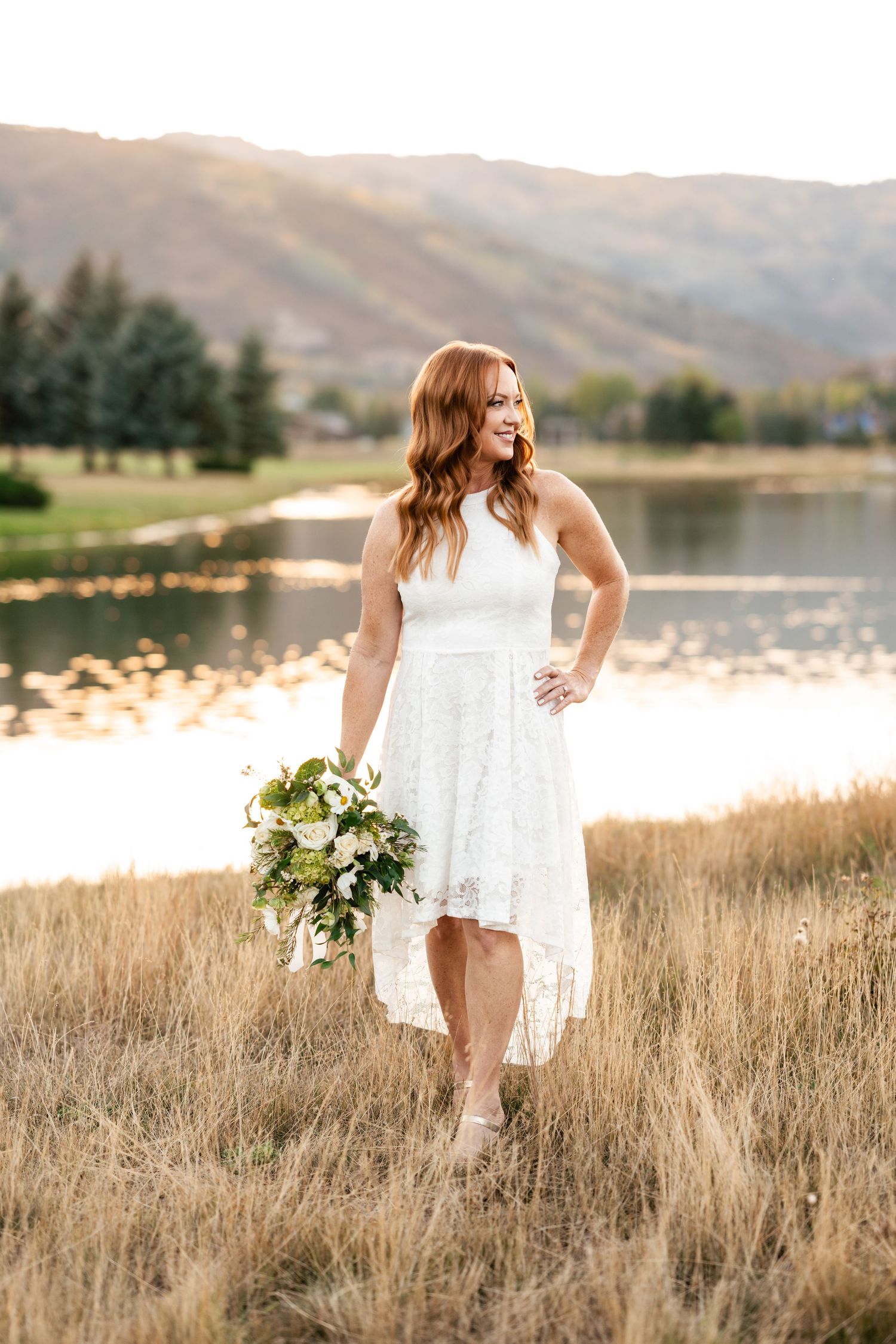 Bride in white dress holds bouquet while standing in golden field by lake.