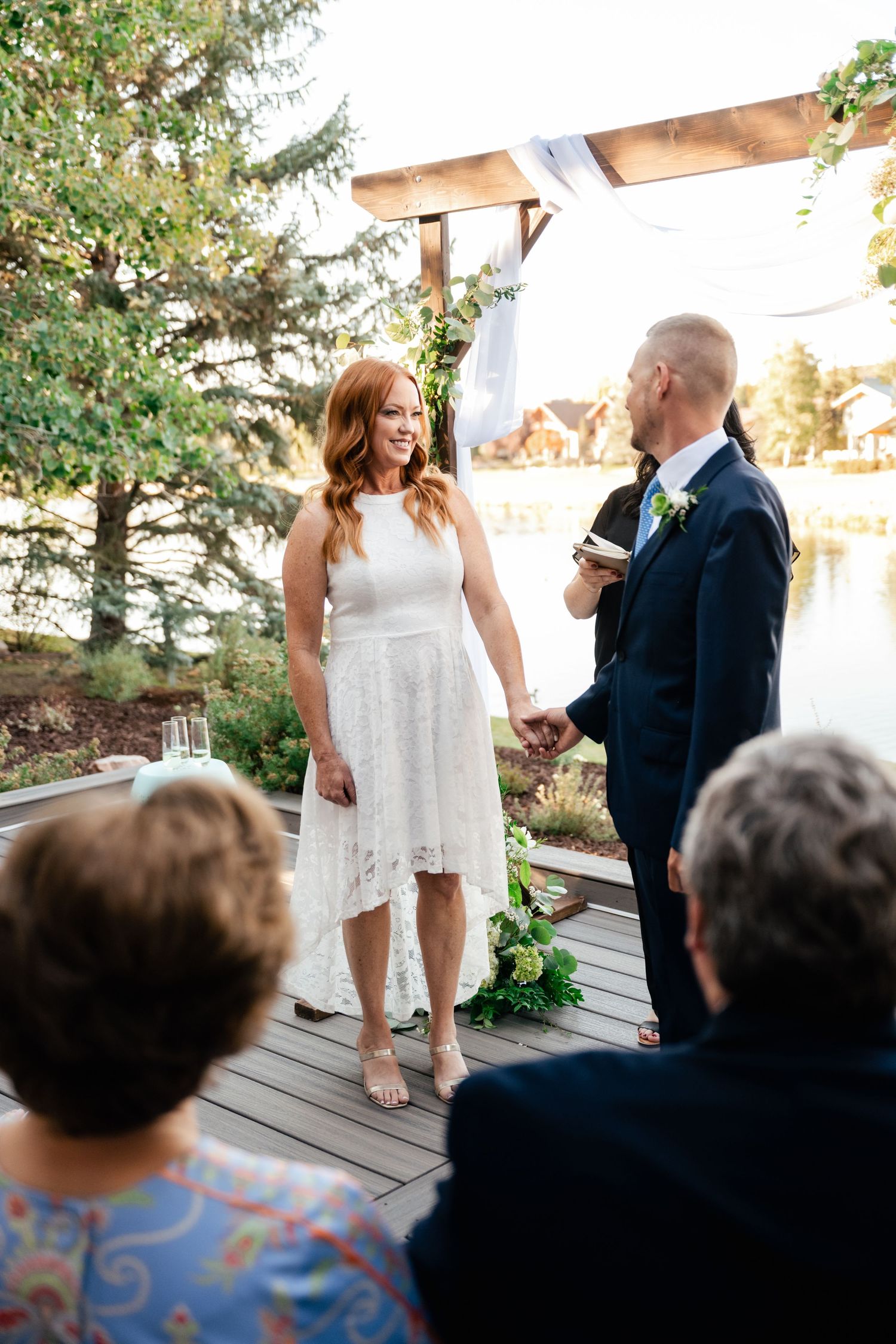 Wedding ceremony on an outdoor deck with white decor and greenery in the background.