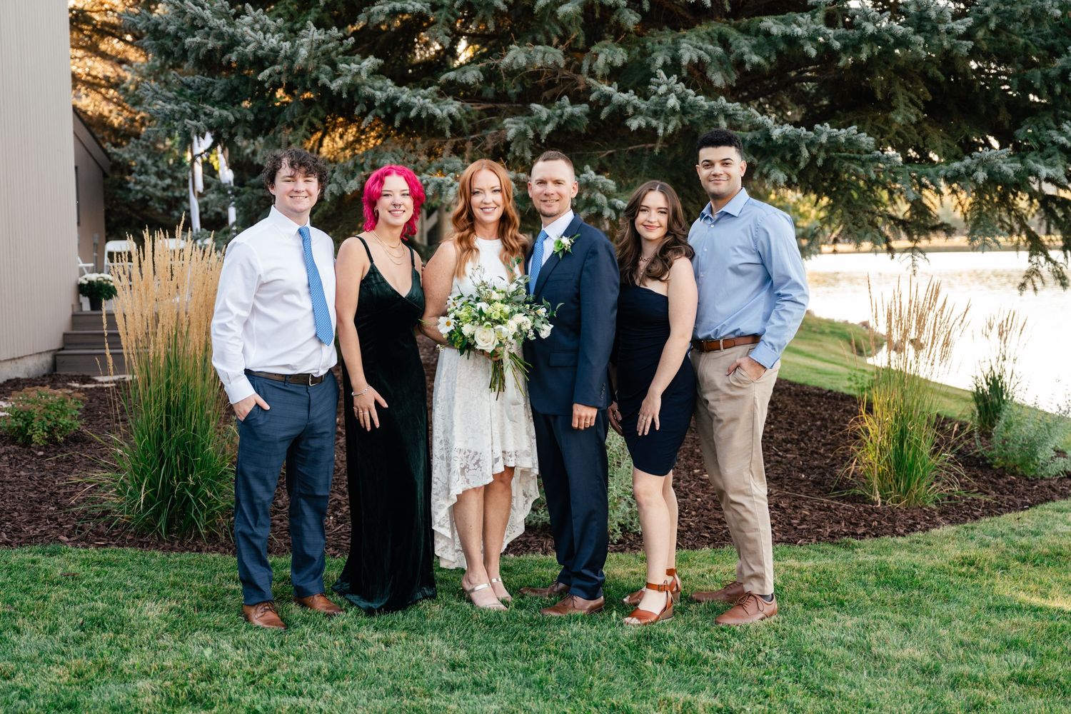 A wedding party stands together on a lawn with blue spruce trees and landscaping behind them.