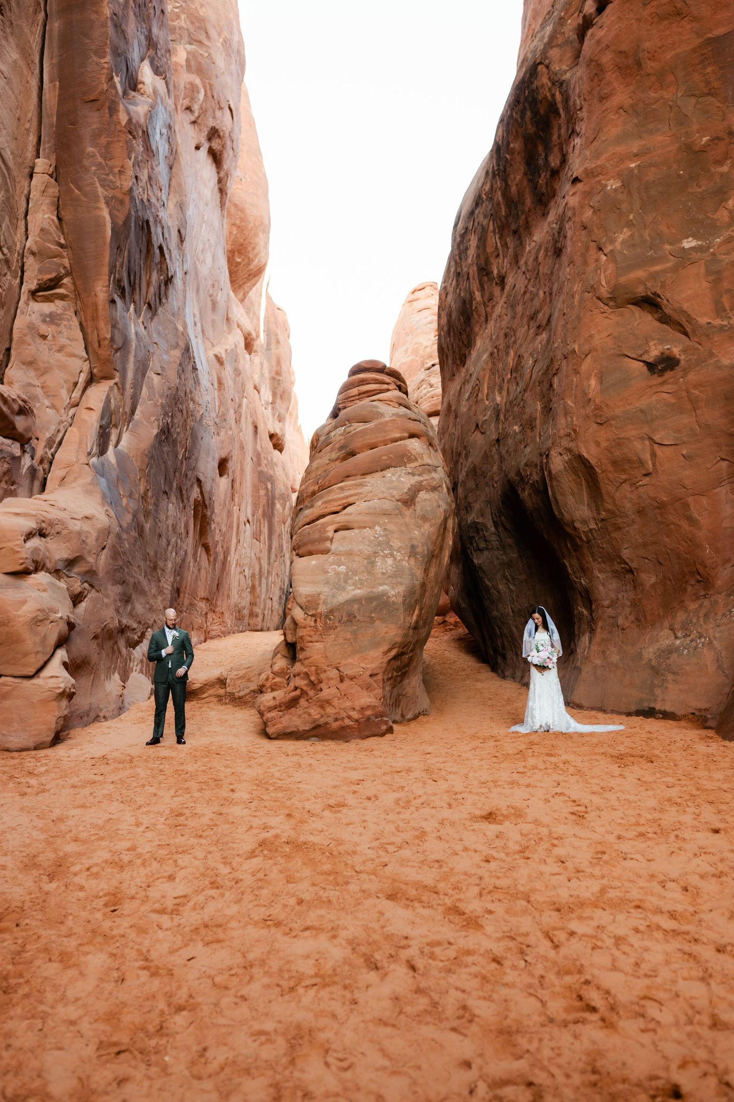 Wedding couple poses between towering red rock formations in a desert landscape.