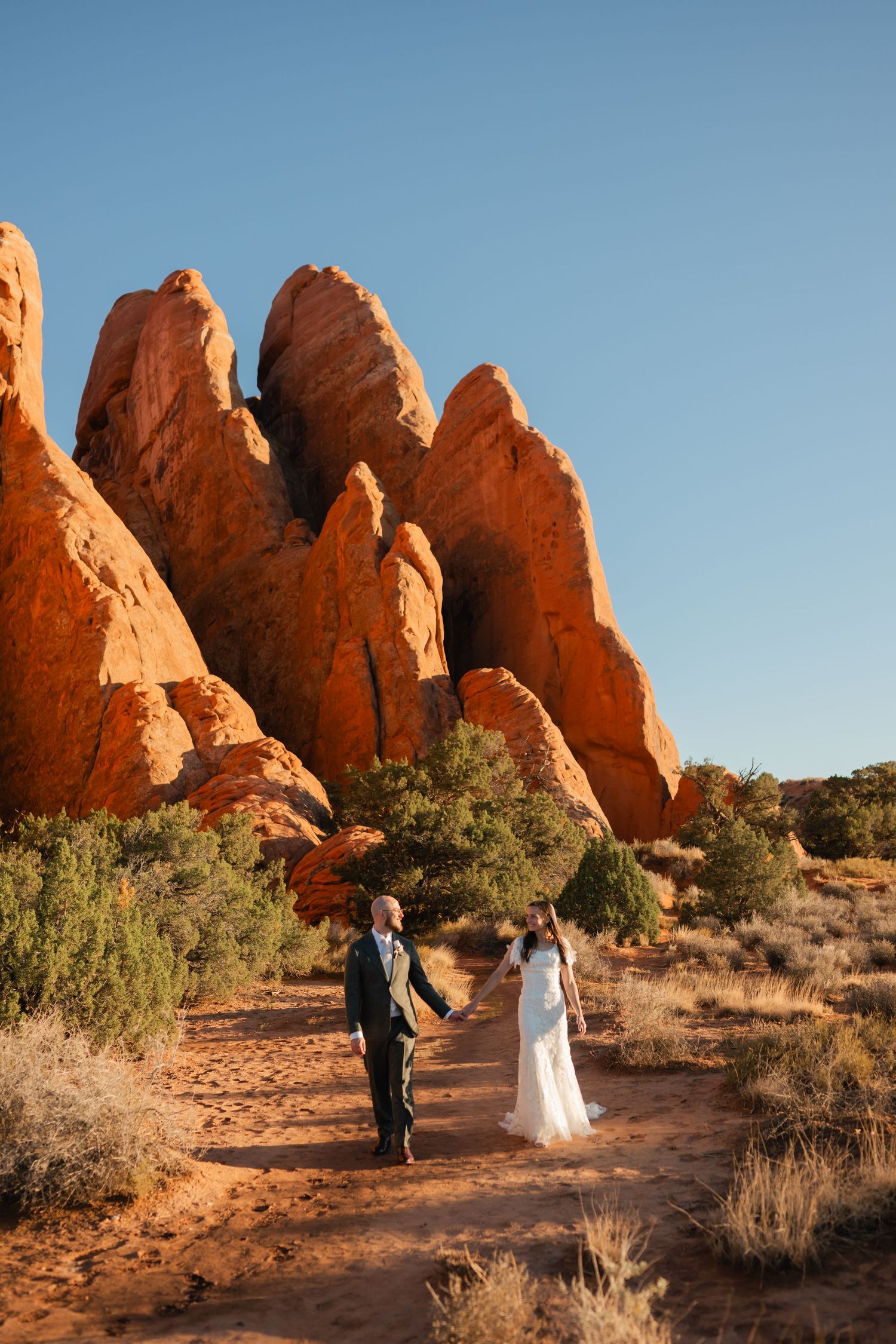 A wedding couple poses on a desert trail with towering red rock formations in Arches National Park.