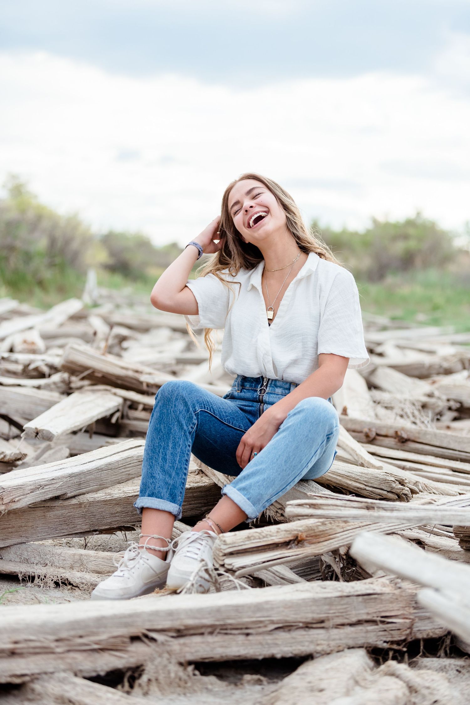 A person in jeans and white shirt laughs joyfully while sitting on driftwood at the beach.