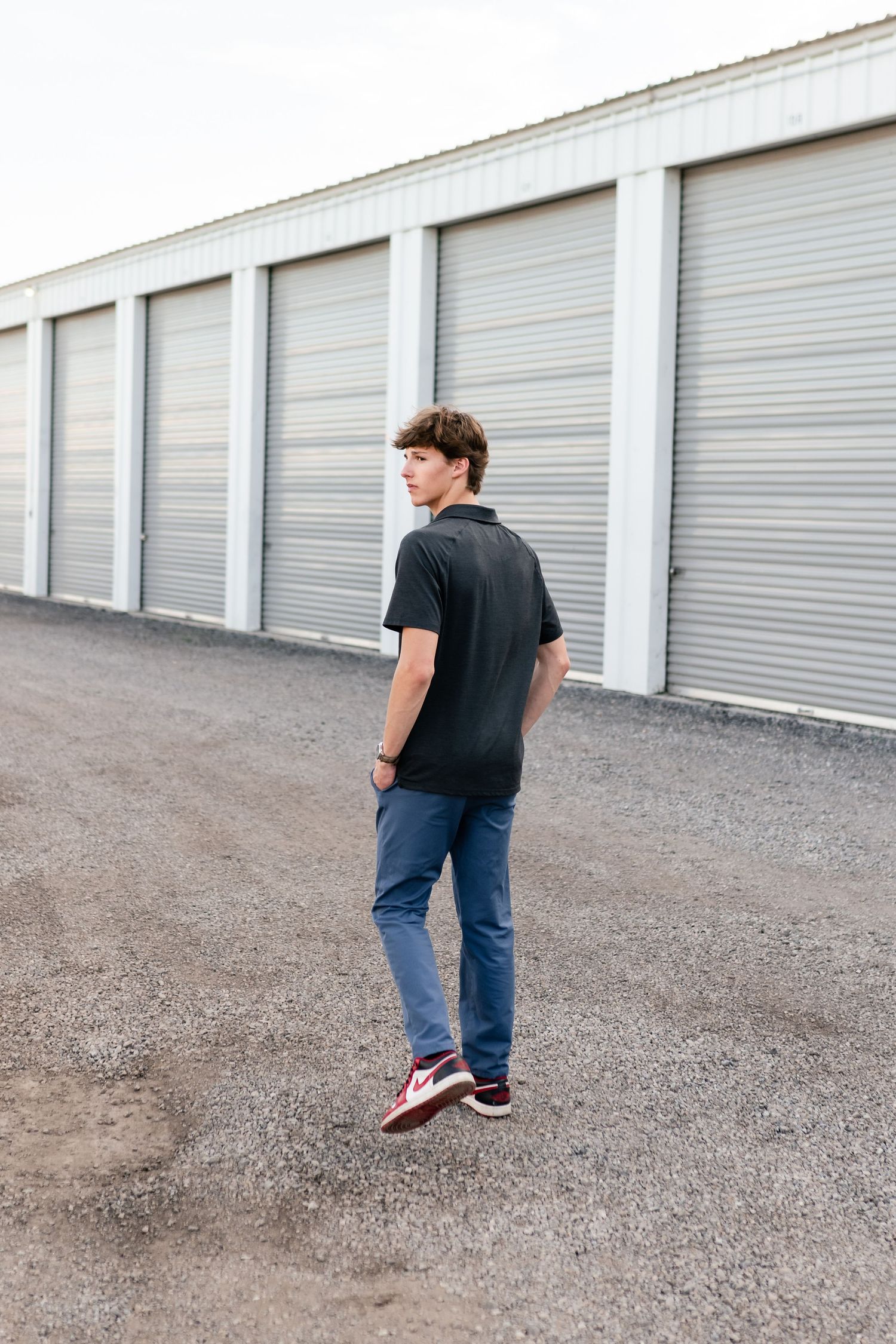 Person in black shirt and jeans walks past white storage units on gravel ground at sunset.