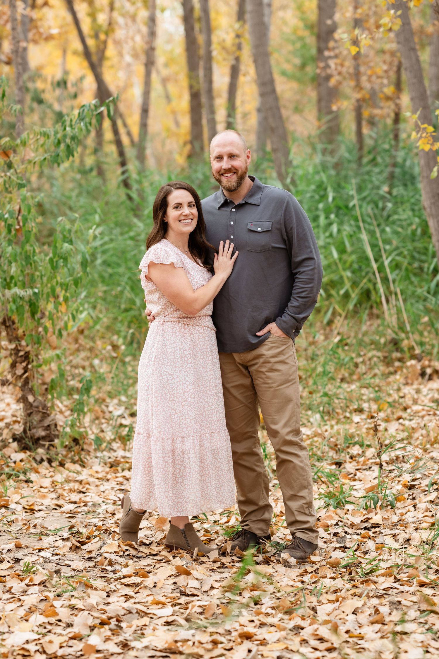 A couple poses together in a scenic autumn forest setting surrounded by fallen leaves and trees.