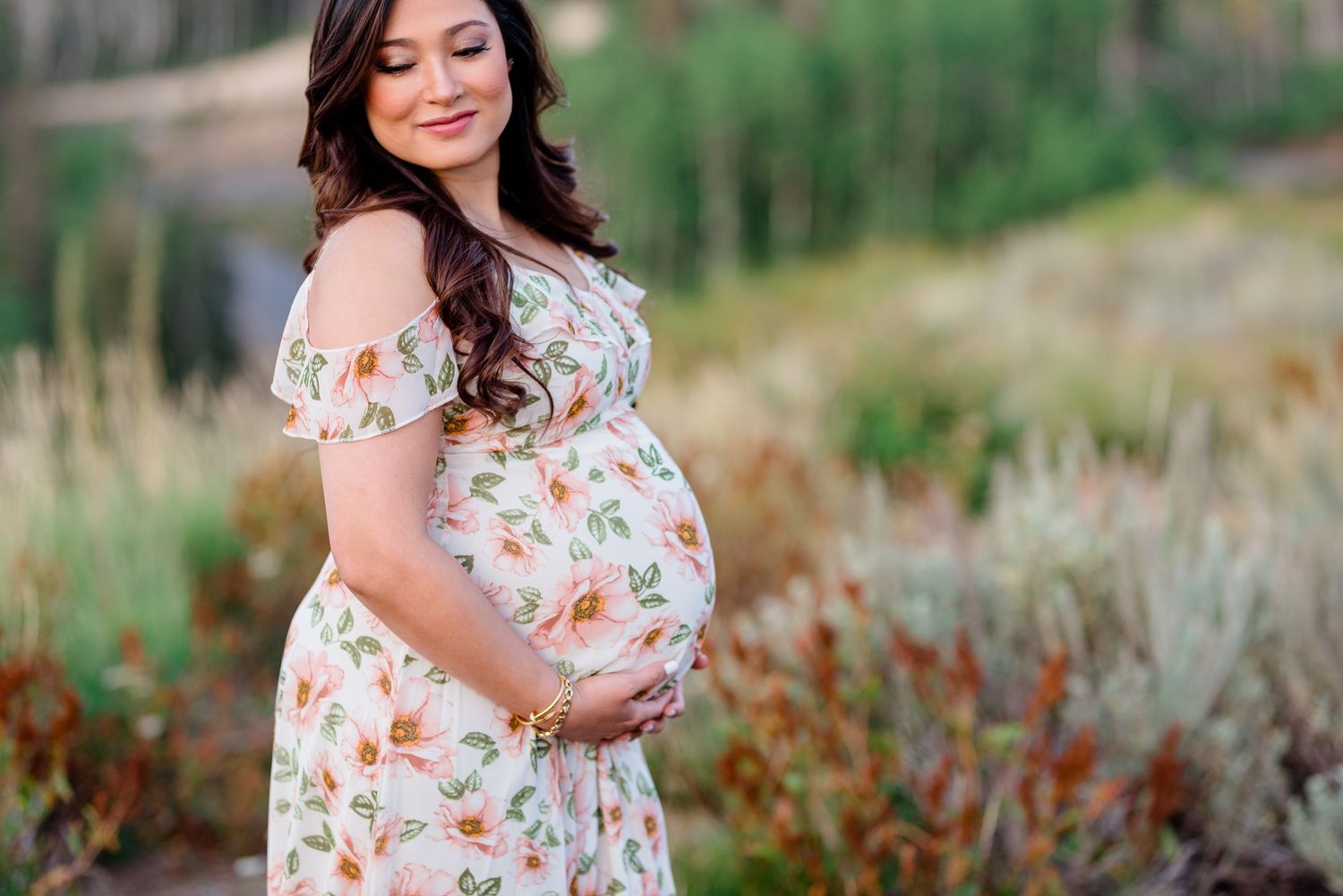 A woman wearing a white floral sundress stands among blooming wildflowers in a scenic mountain landscape.