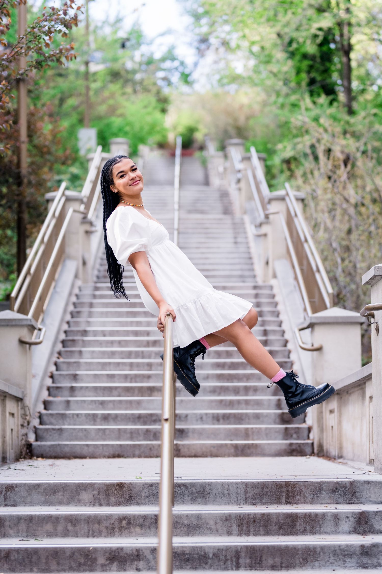 A person in a white dress and combat boots playfully leans on a handrail of an outdoor concrete staircase.