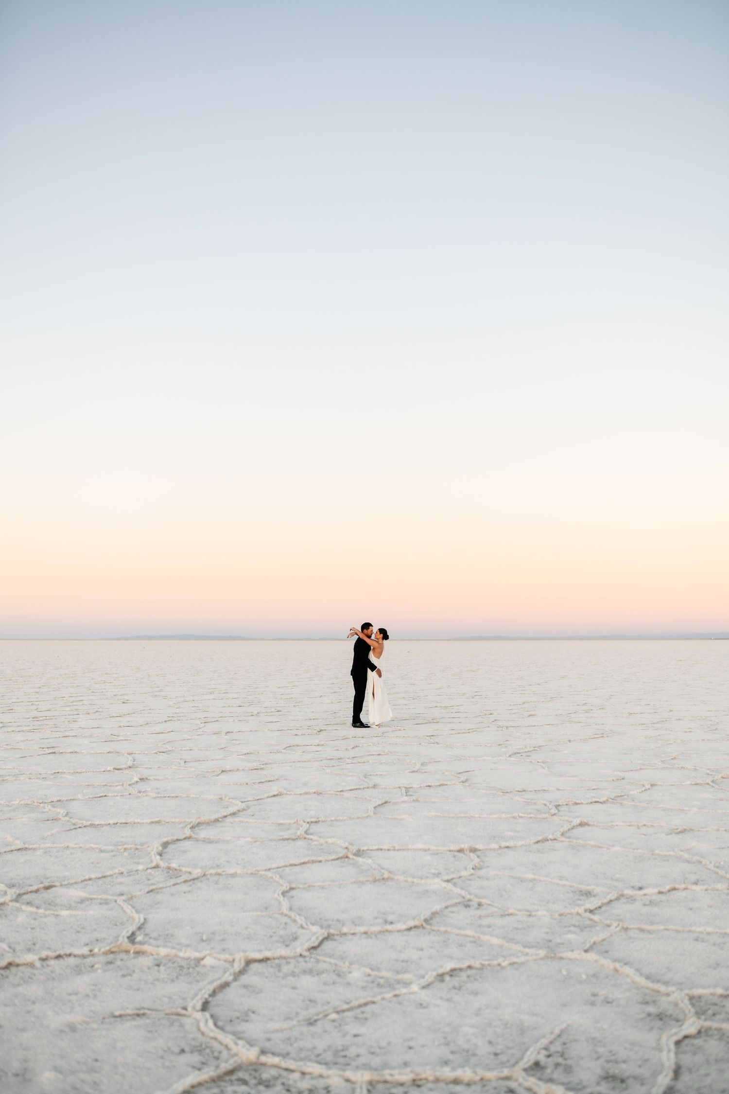 Utah Salt Flats Photographer. Two distant figures stand on vast white salt flats against a pastel sunset sky.