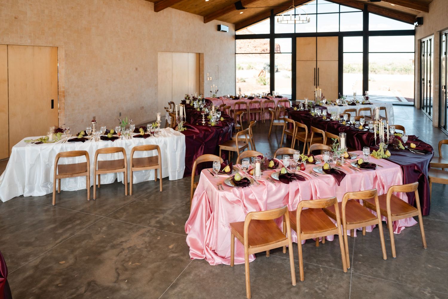 Elegant wedding reception hall with pink and burgundy tablecloths, wooden chairs, floral centerpieces, and large windows.