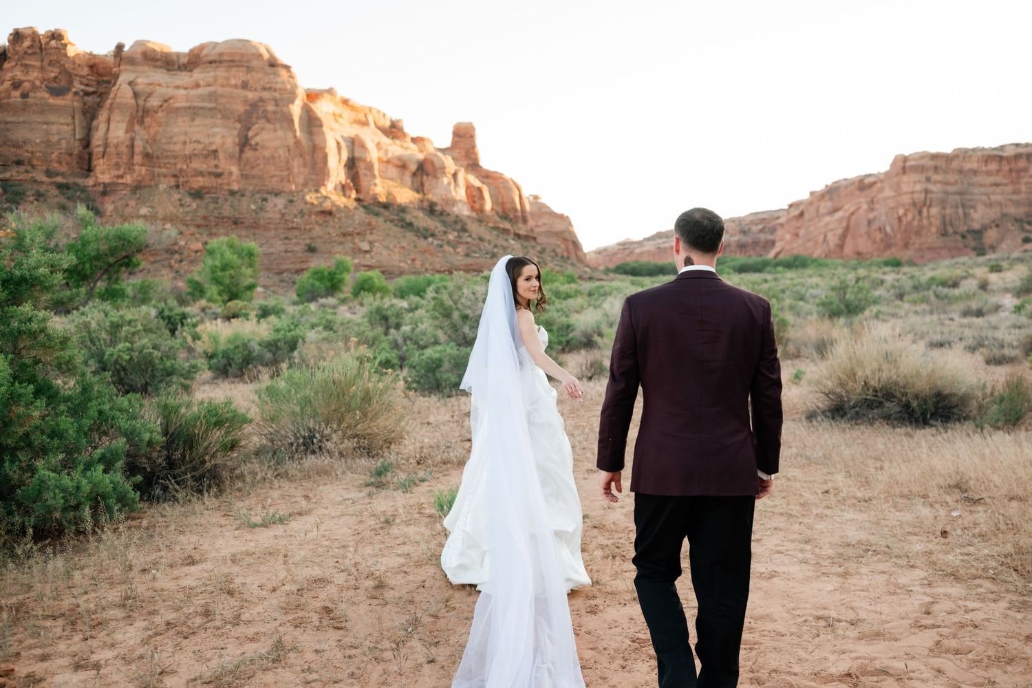 Bride and groom walking hand in hand through a scenic desert landscape with red rock formations.