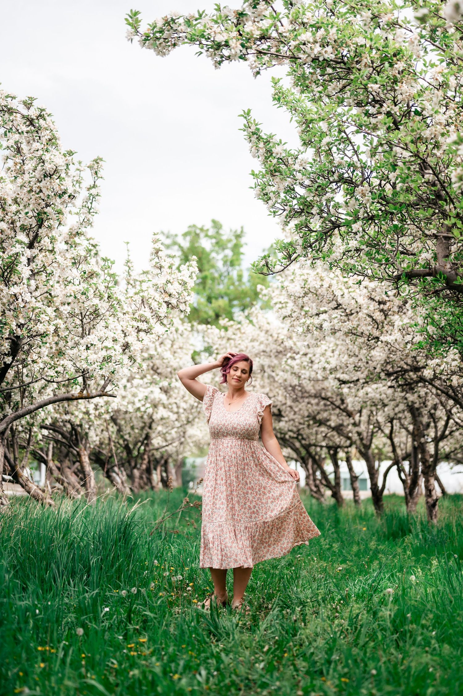 A dreamy spring scene with blooming cherry trees and a person in a flowy polka dot dress standing in green grass.
