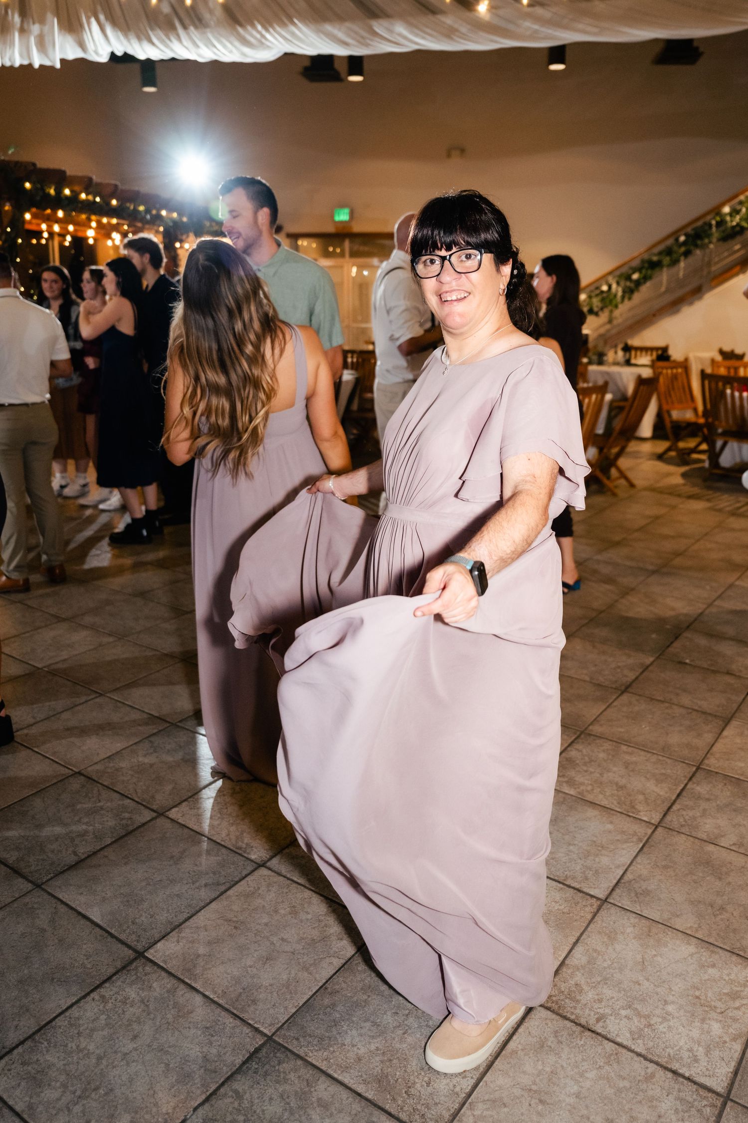 Wedding guest in a flowy mauve bridesmaid dress dancing at a reception with string lights and draped ceiling decor.