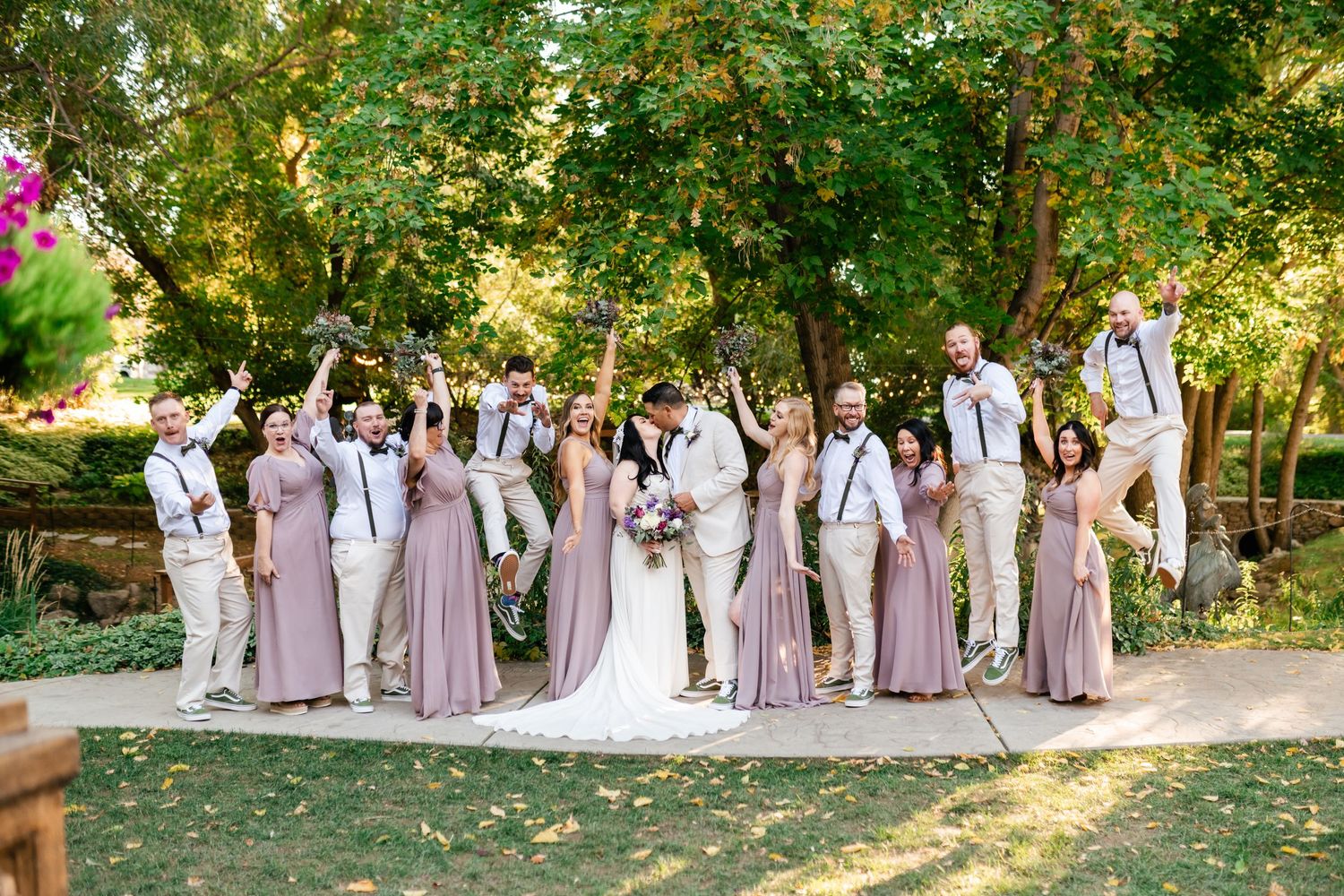 Wedding party poses in coordinated blush and white outfits underneath lush green trees in a garden setting.