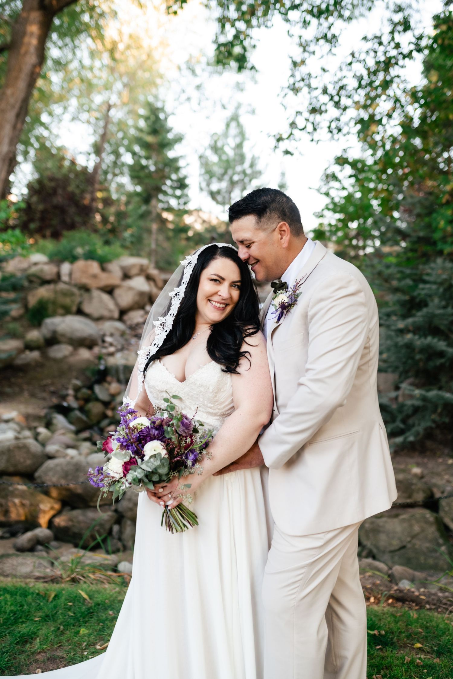A joyful couple shares an intimate moment at their outdoor wedding ceremony with a beautiful purple floral bouquet in hand.