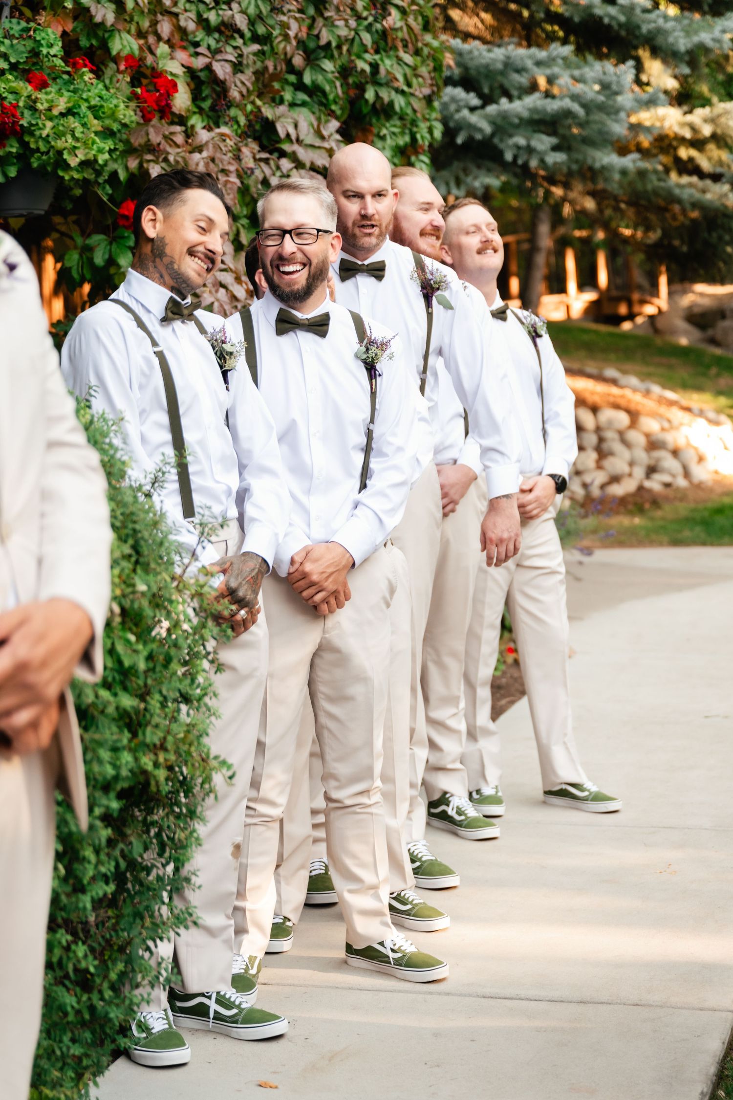 Groomsmen in white shirts and khaki pants laugh together while wearing matching green sneakers at an outdoor wedding venue.