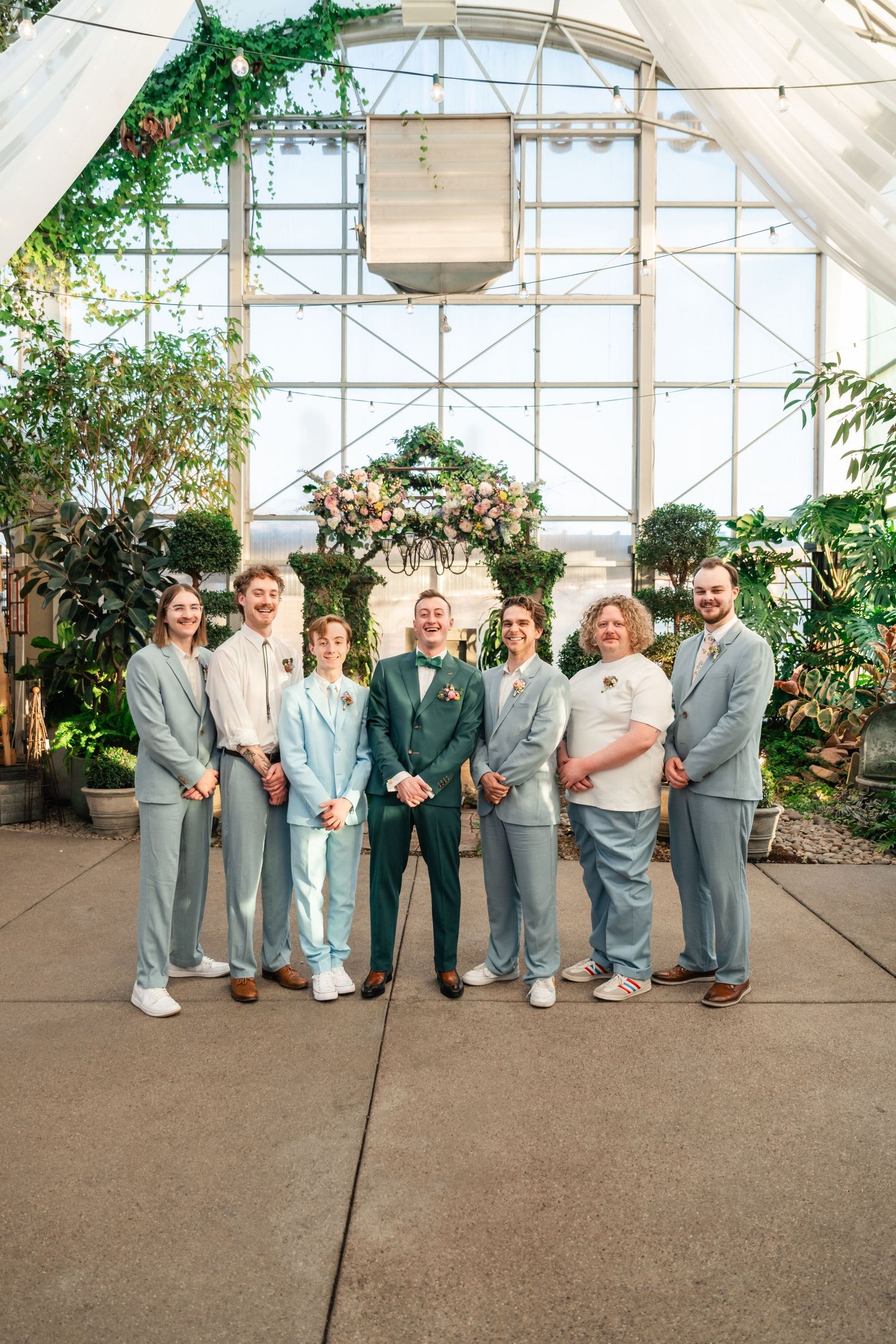 Wedding party in coordinated light grey suits and dresses posed in front of a greenhouse with hanging greenery.