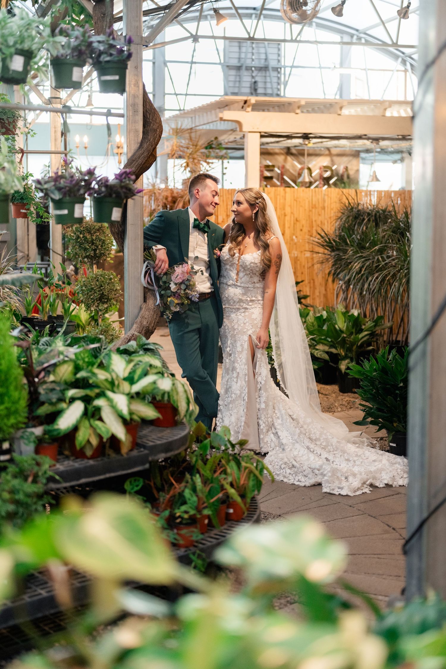 A romantic wedding couple stands surrounded by lush greenery and potted plants in a sunlit greenhouse conservatory.