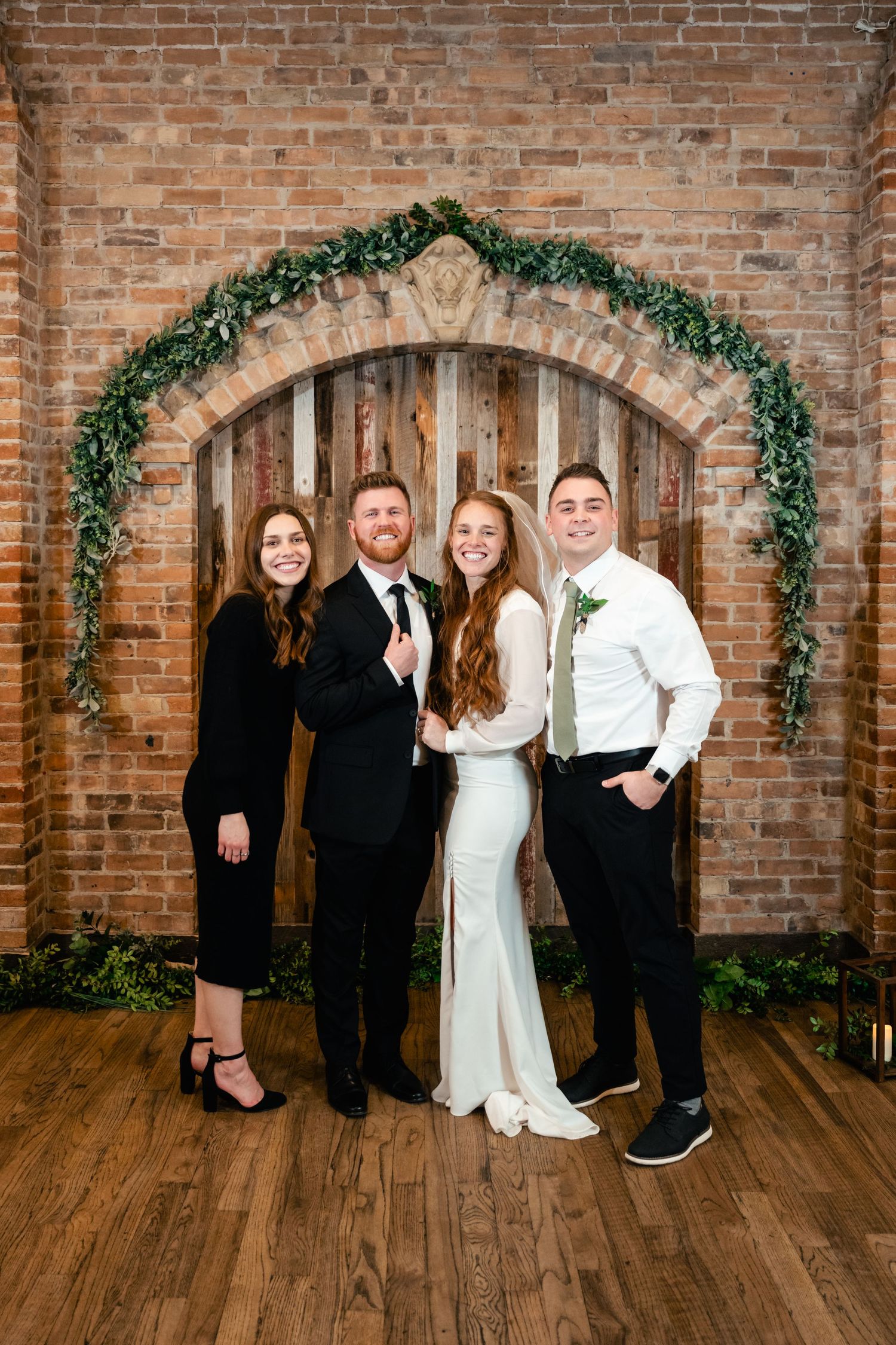 Wedding group poses under elegant brick archway decorated with greenery garland at indoor venue.
