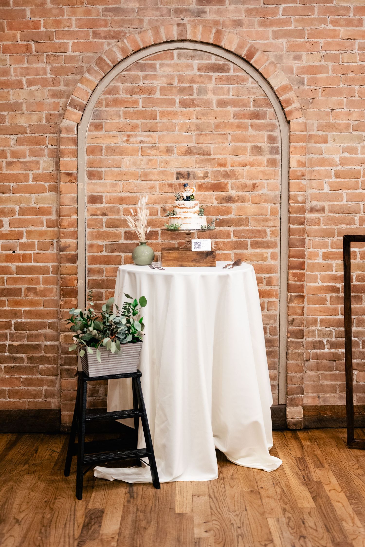 A three-tier white wedding cake displayed on a covered table beneath a brick archway with greenery decor and wooden floors.