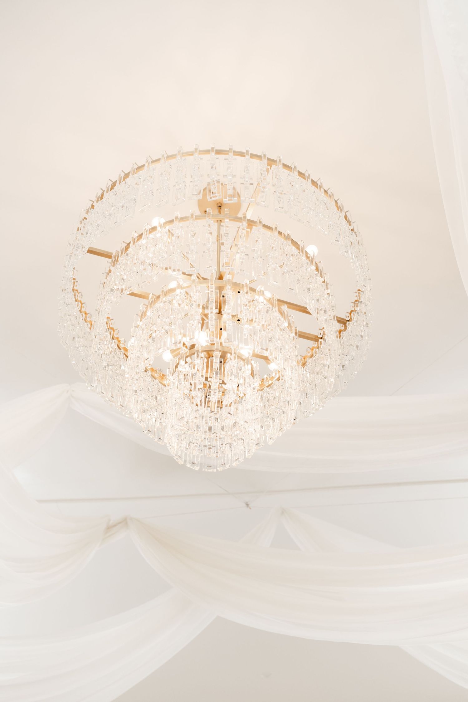 Elegant crystal chandelier with cascading tiers hanging from draped white fabric ceiling decor. Moments on Main by Utah Wedding Photographer