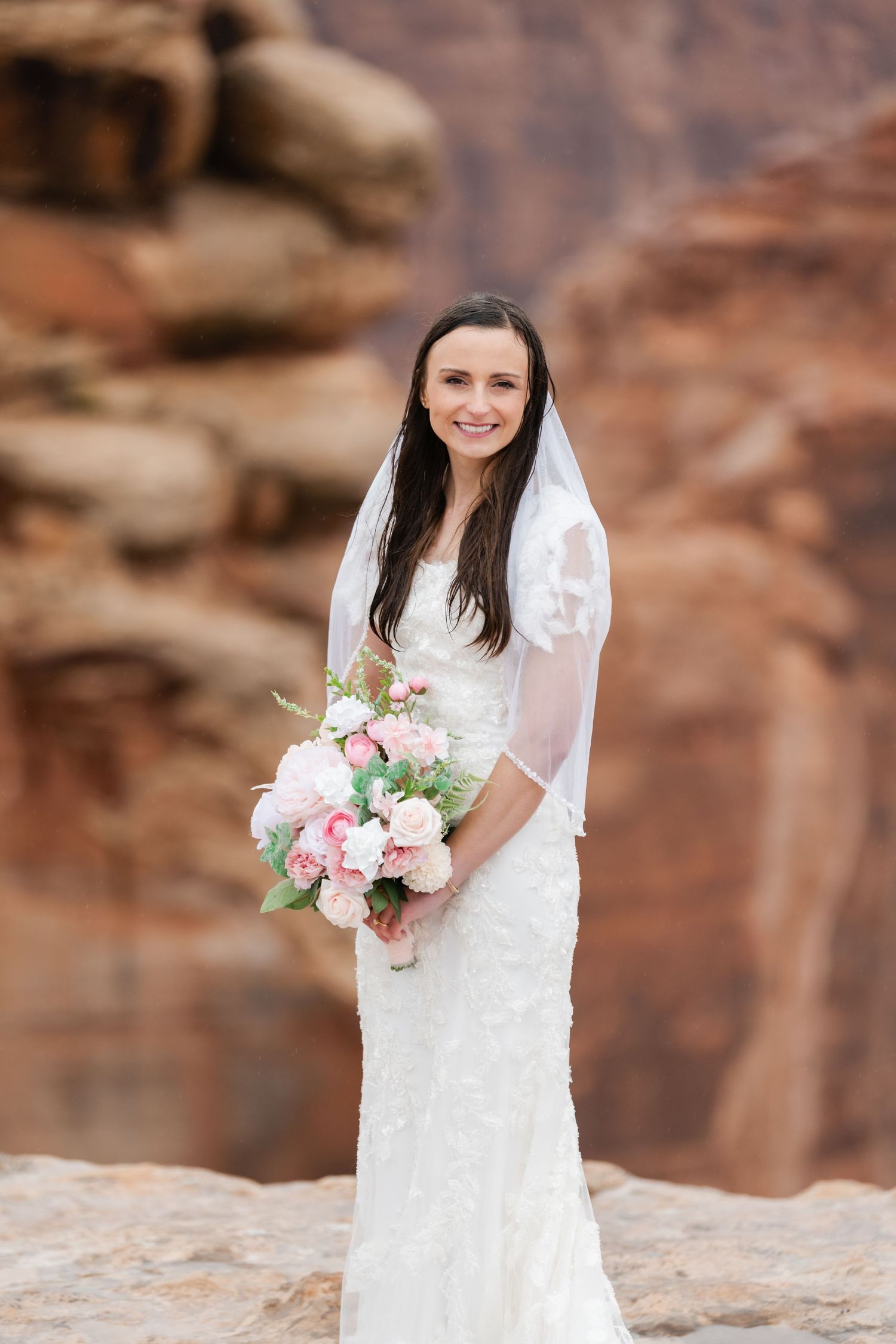 A bride poses with her bouquet of pink and white flowers against red rock canyon walls.