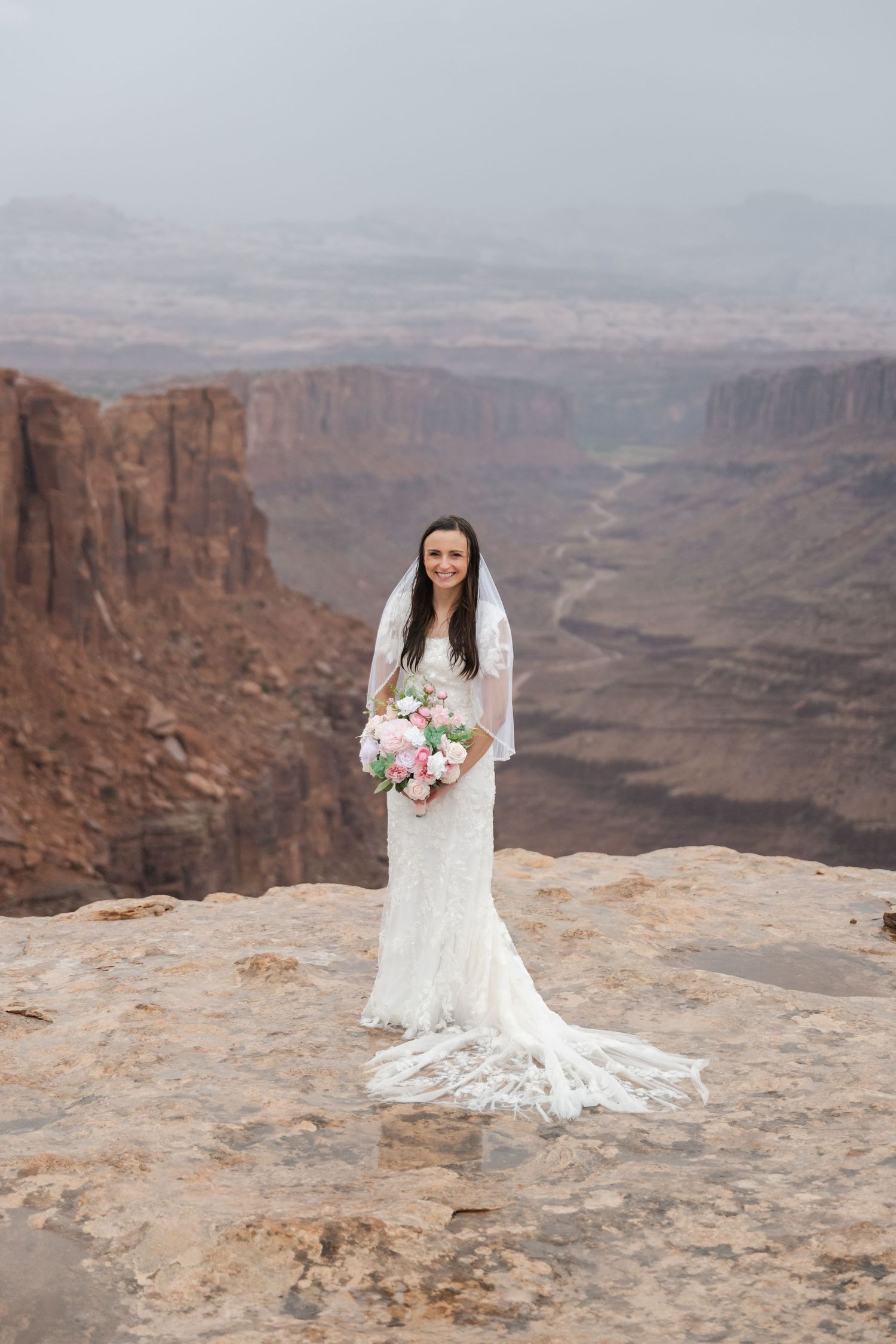 A bride in a lace wedding dress poses on the edge of a majestic desert canyon overlook.