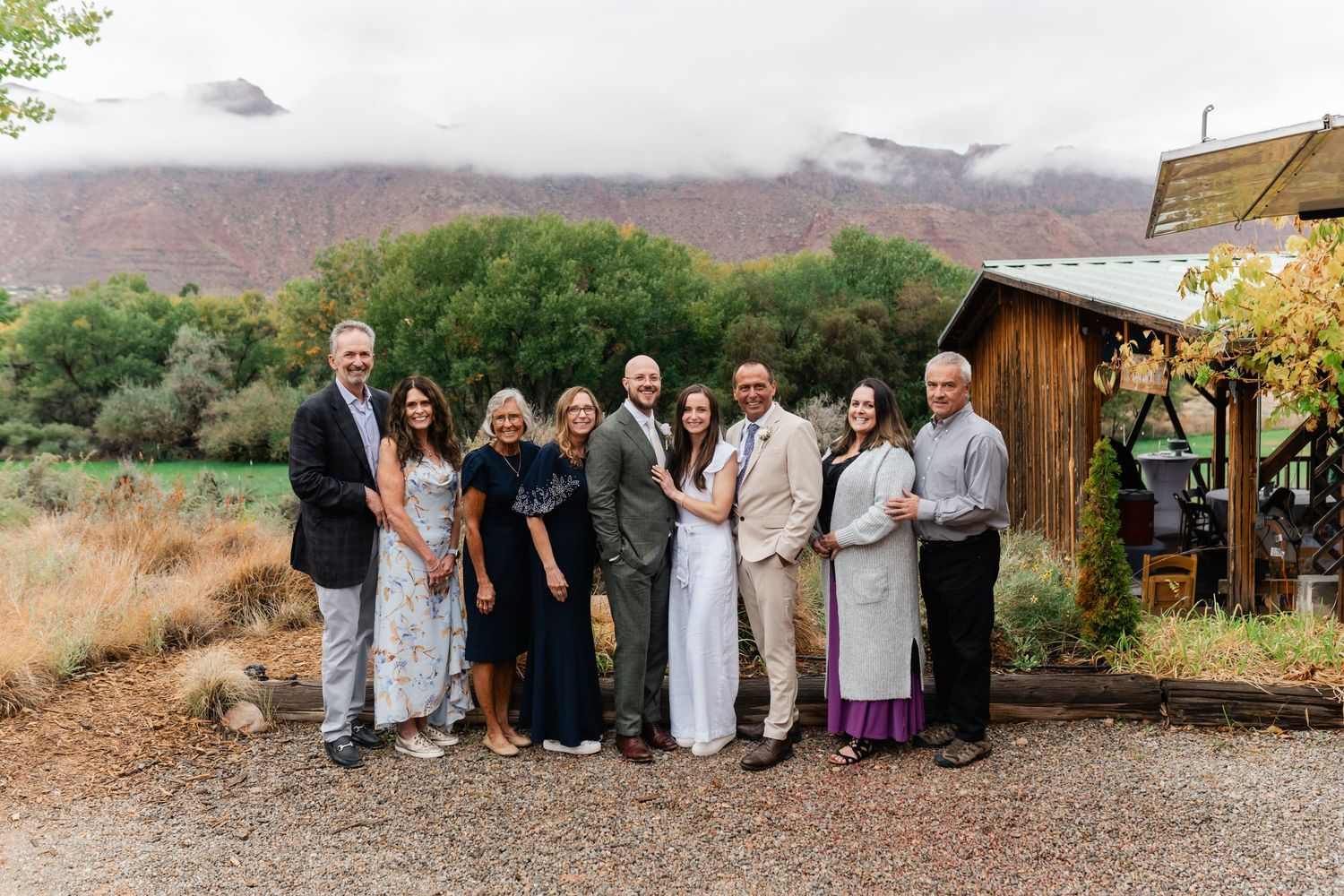 Wedding celebration group photo taken outside rustic wooden buildings with desert mountains in background.