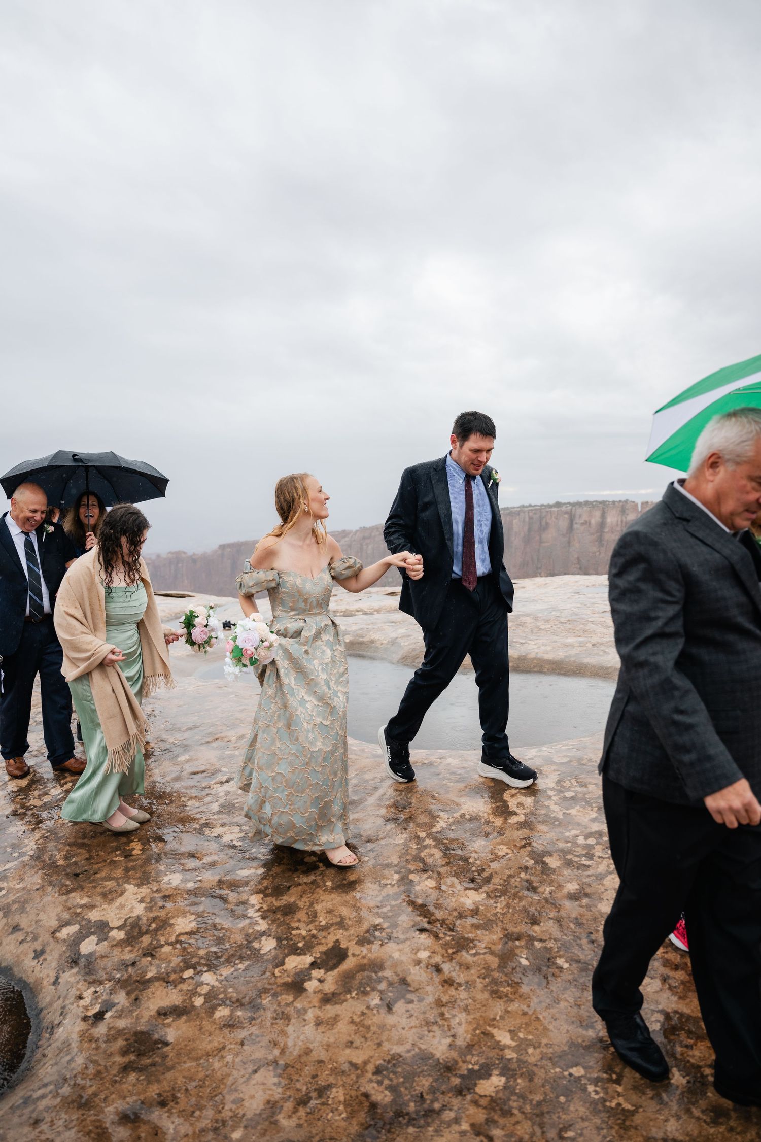 Wedding party walking on rocky terrain during overcast day with umbrellas and formal attire.
