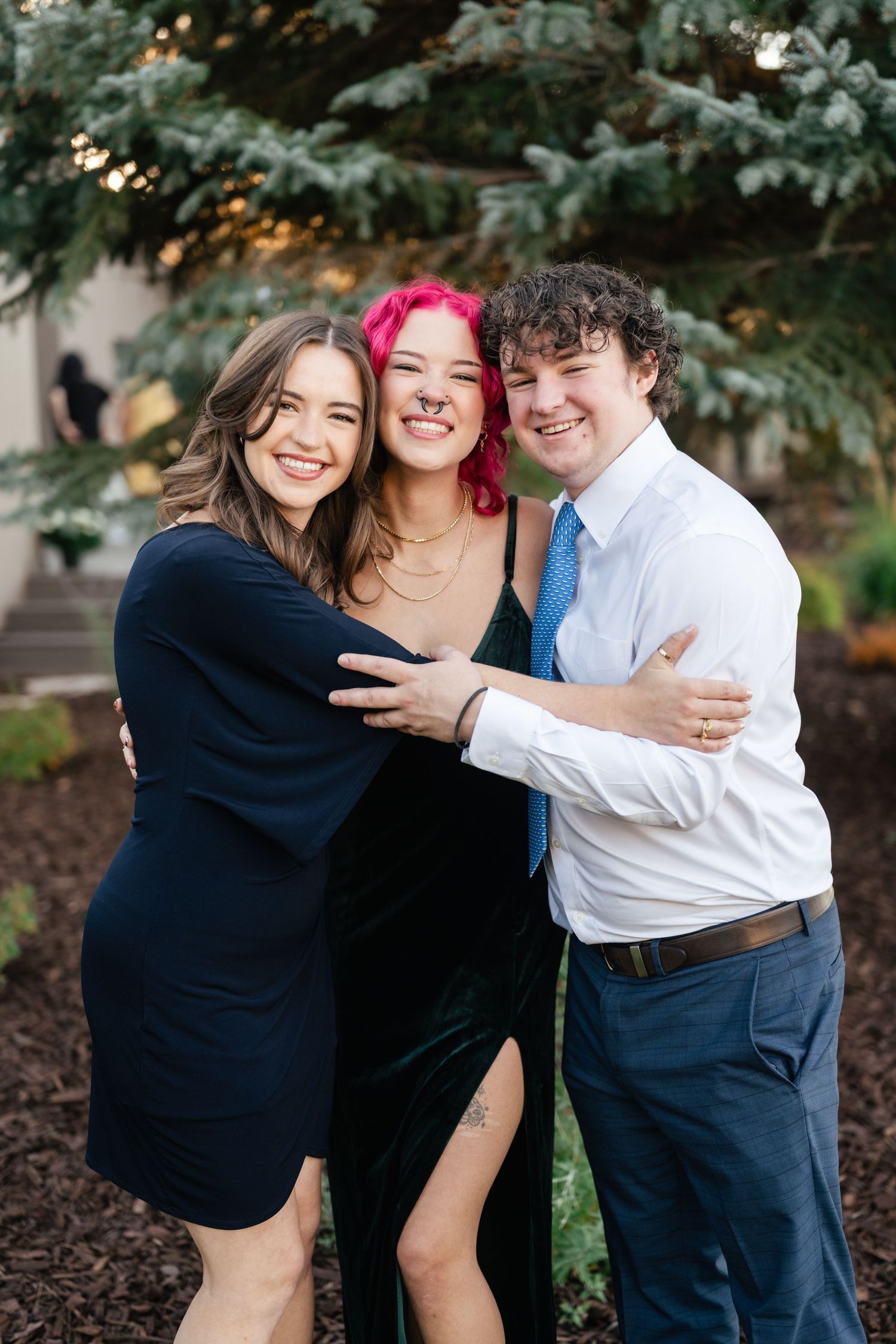 Three people embracing and laughing together outdoors near evergreen trees.