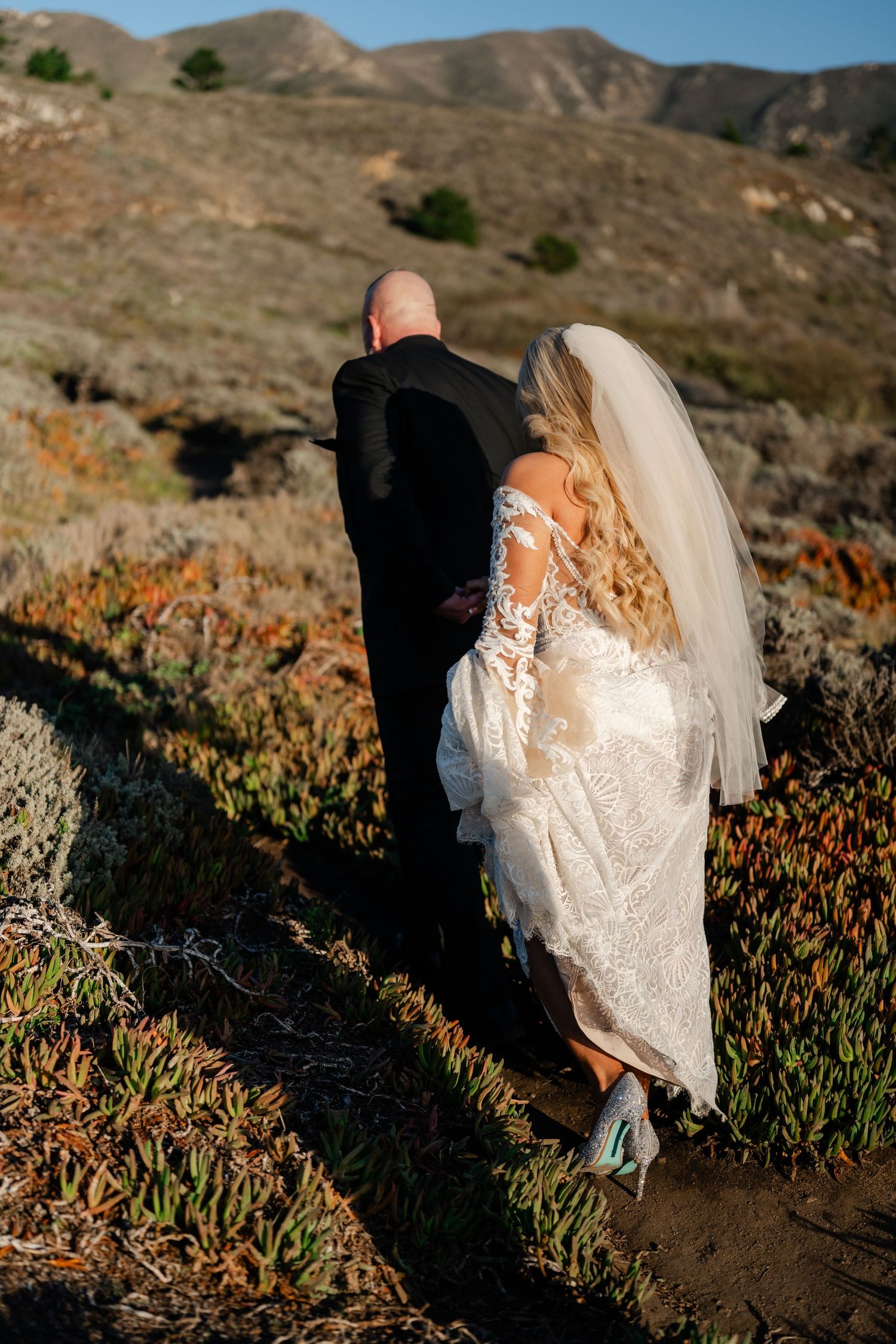A couple walks along a scenic mountain trail, the bride in a lace gown with flowing veil against rugged terrain.