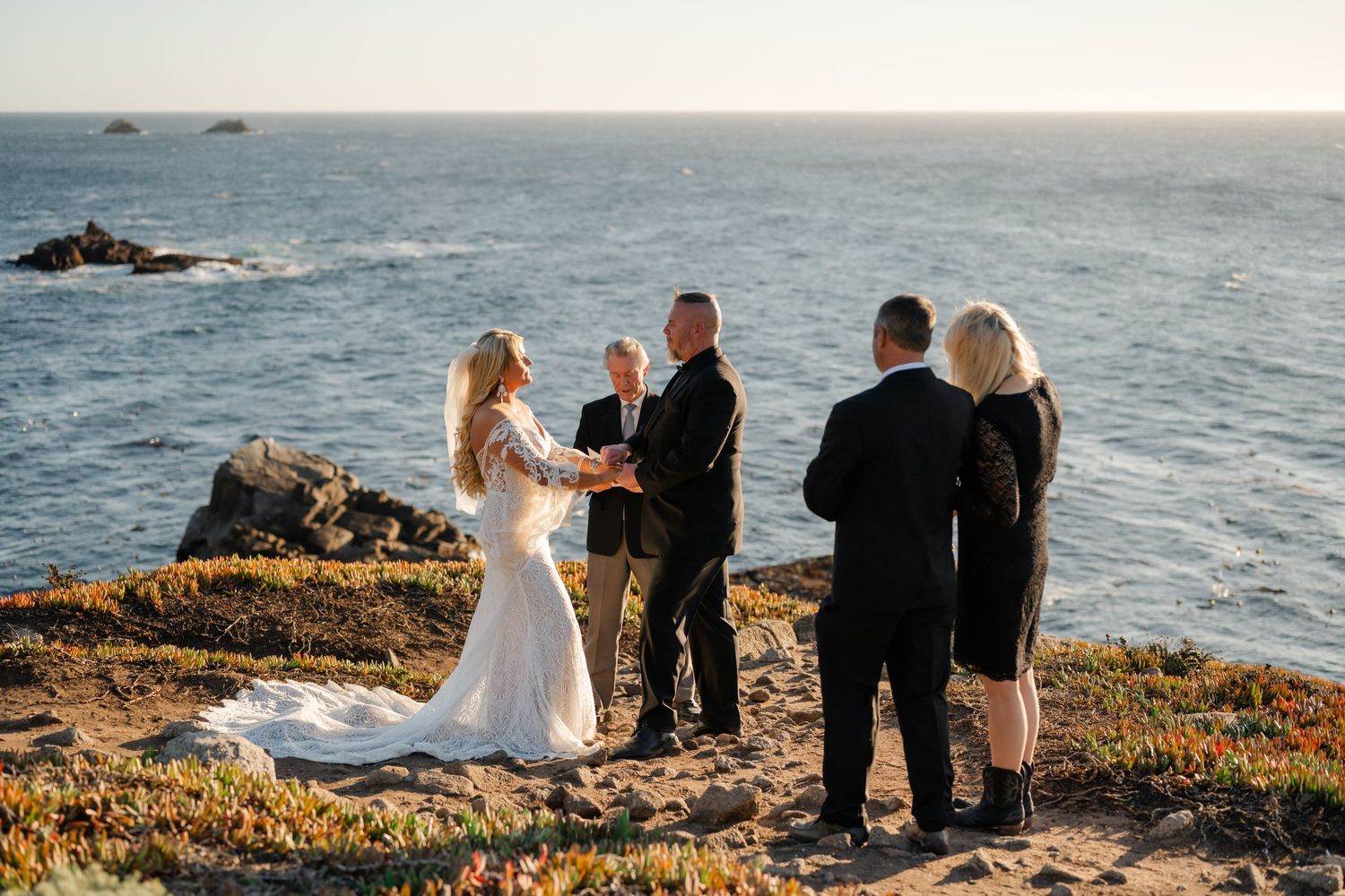 An intimate wedding ceremony unfolds on a rugged oceanside cliff with waves crashing below.