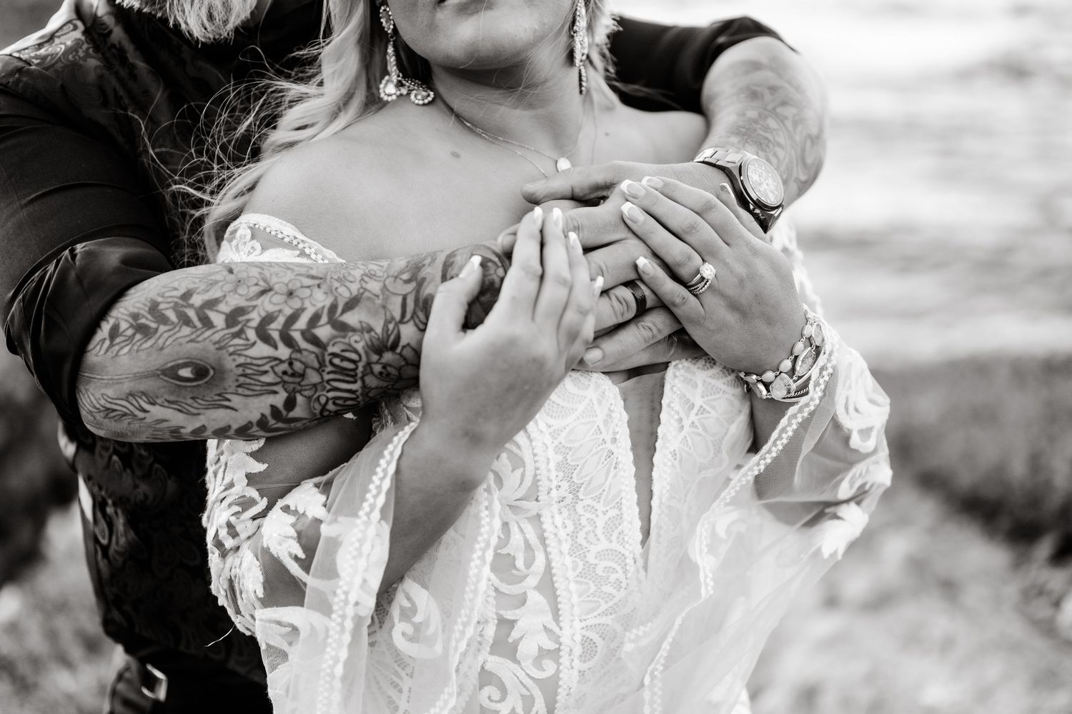 Close-up black and white photo showing tattooed hands embracing in romantic moment against natural backdrop.