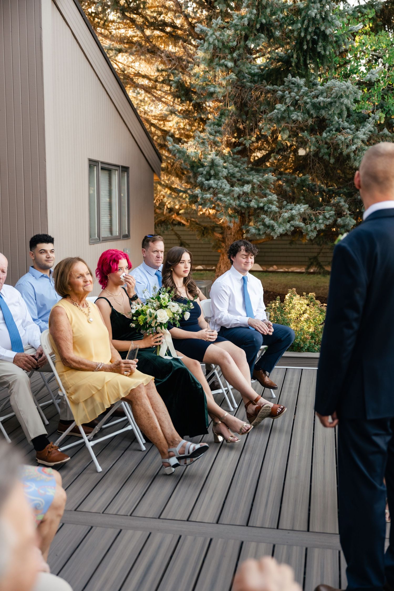 Wedding guests seated in rows on a wooden deck during an outdoor ceremony with evergreen trees in the background.