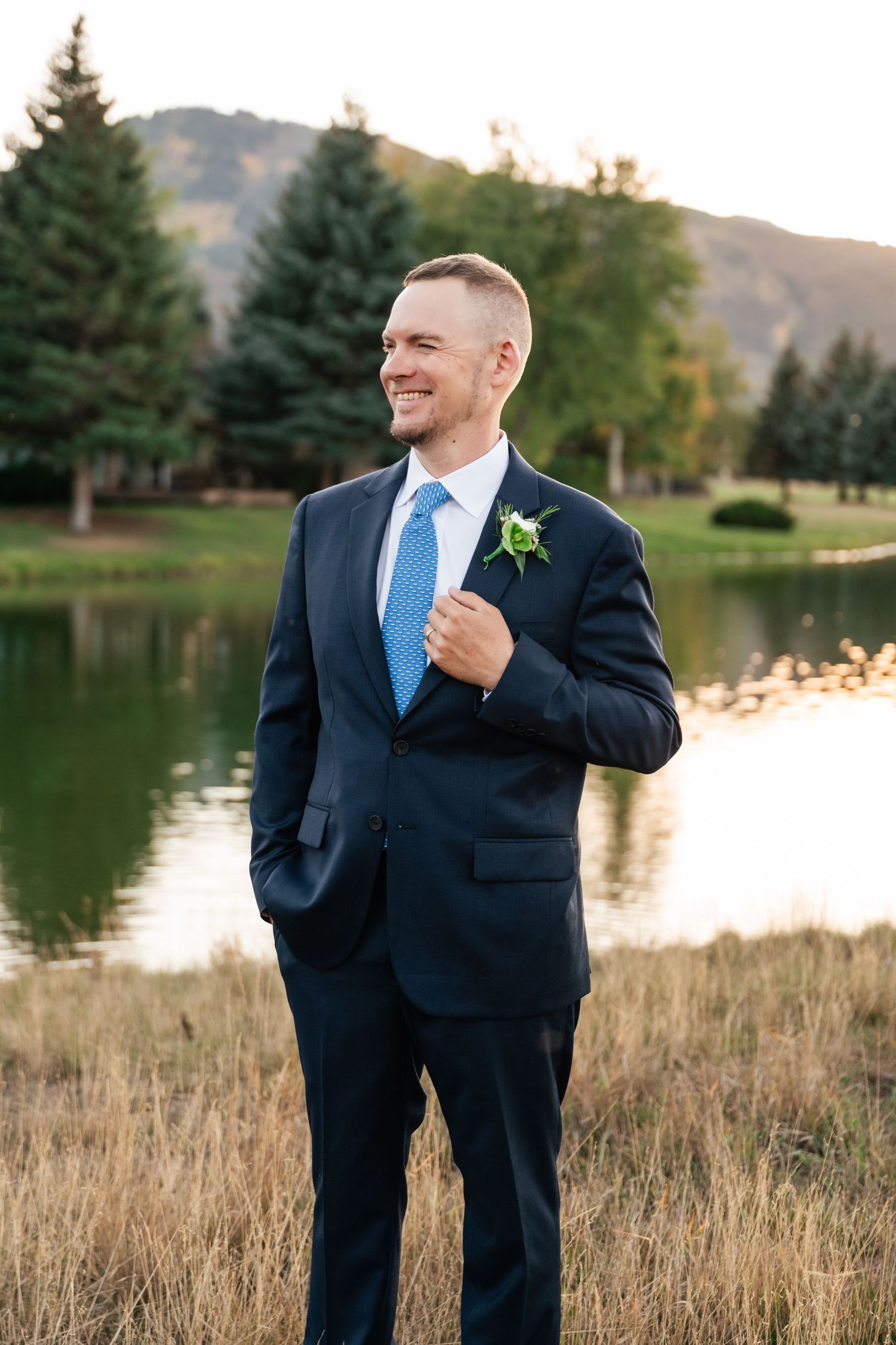 A groom in a navy suit and blue tie poses outdoors near a lake with mountains and evergreen trees in the background.